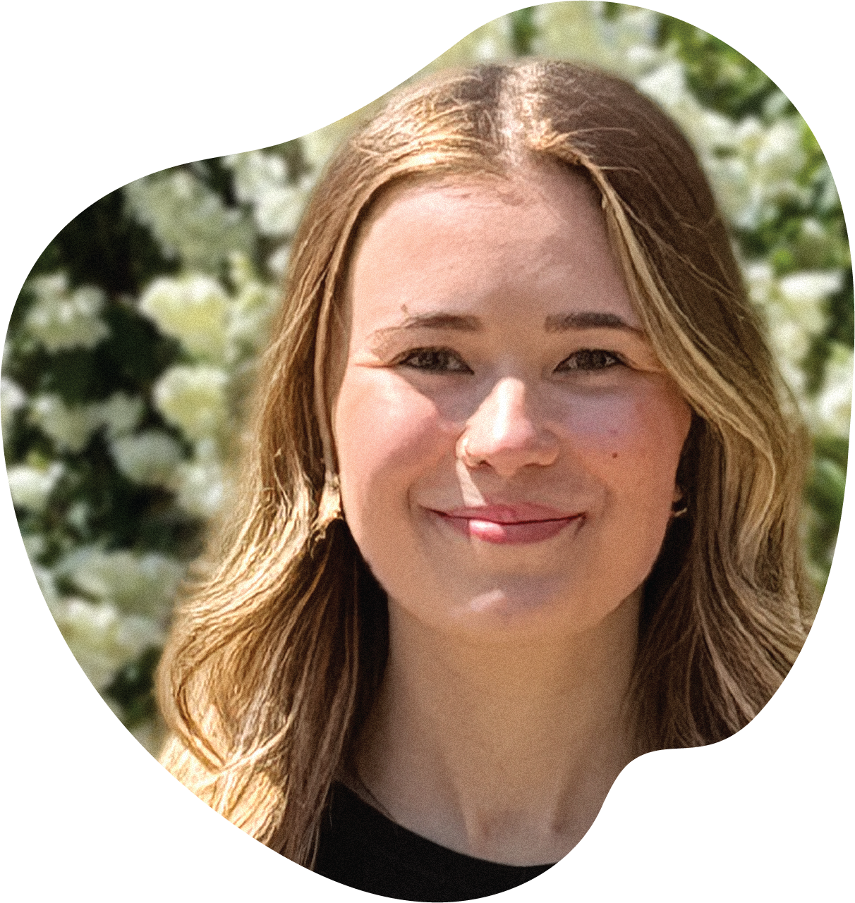 Jill Gerlof is smiling with closed lips. She has blonde hair. behind her is out of focus bush with white flowers. The photo is cropped into the loose shape of an apple.