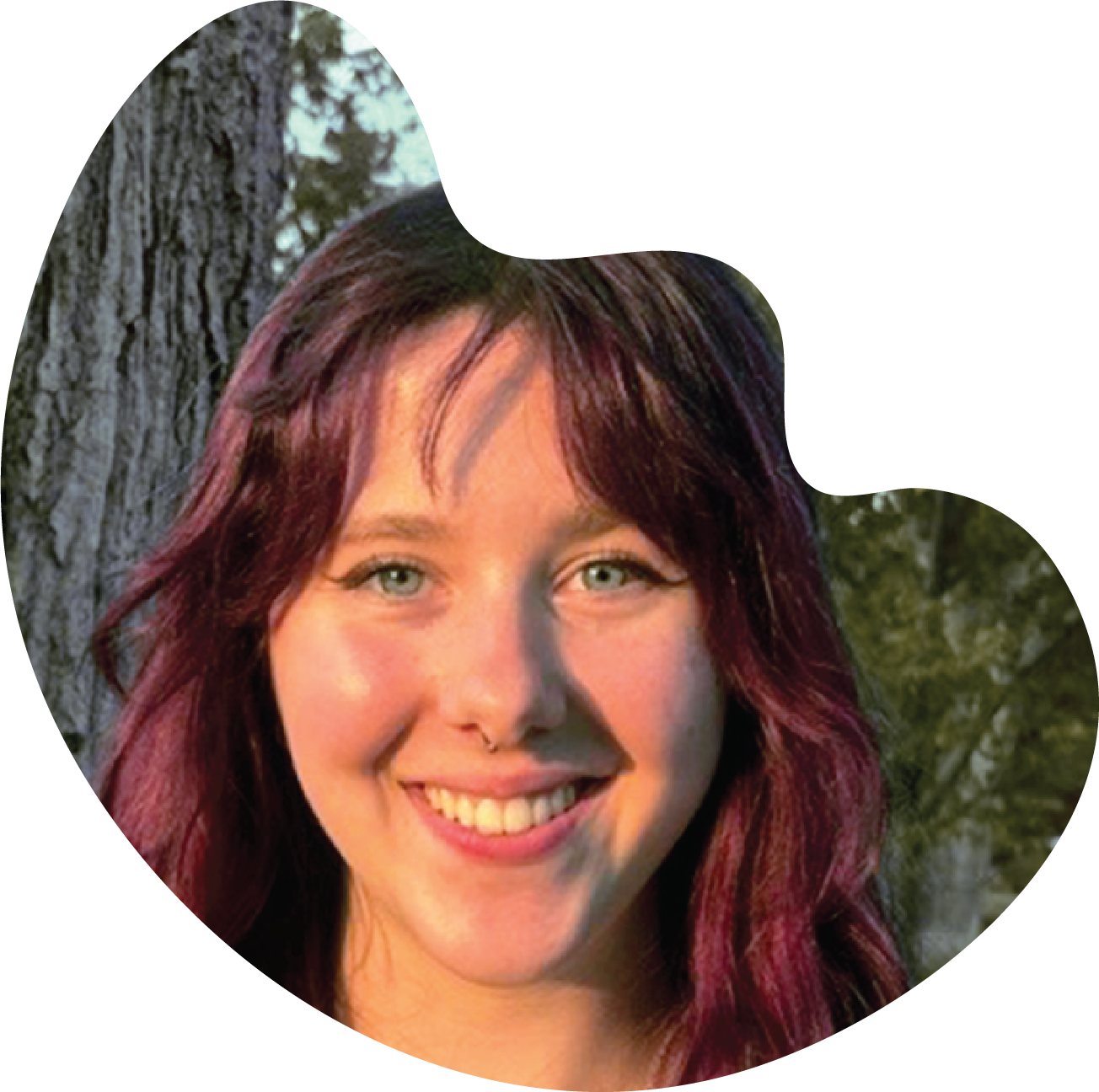 Maggie Myrne is grinning. She has died aubergine hair and is wearing a septum peircing. behind her is out of focus poplar tree. The photo is cropped into the loose shape of a bloom.