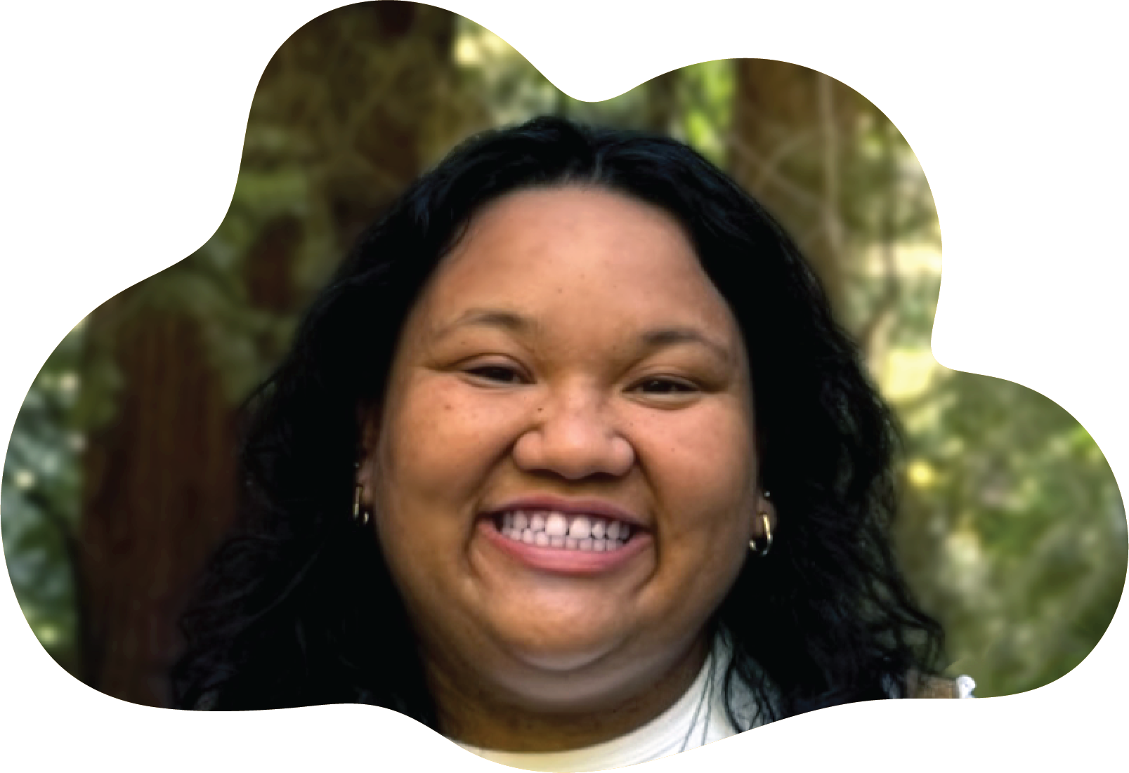 Jenna Nombre looks happy with a bright grin. She has dark hair, small gold hoop earrings. there is out of focus wild parkland in the background. The photo is cropped into the loose shape of a cloud.