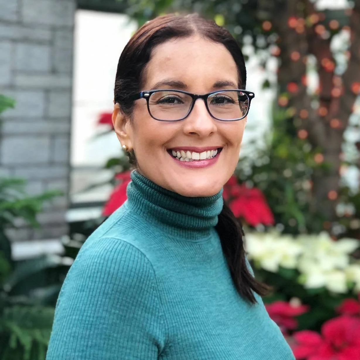 A smiling woman with glasses and dark hair in a ponytail, wearing a teal turtleneck, standing outdoors with colorful flowers and greenery in the background.