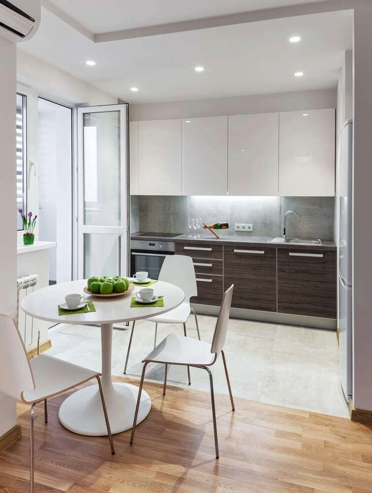 Modern kitchen with white and wood cabinetry, a round dining table with four chairs, green apples and cups on the table, a small balcony door, and a window with purple flowers on the windowsill.