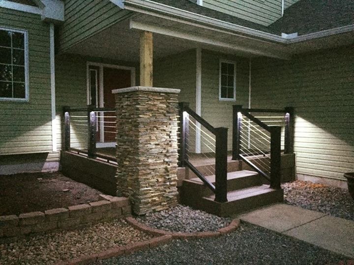 A small outdoor staircase leading to a front porch of a house, with stone columns on either side, black railings with white cables, and gravel along the stairs, at night.