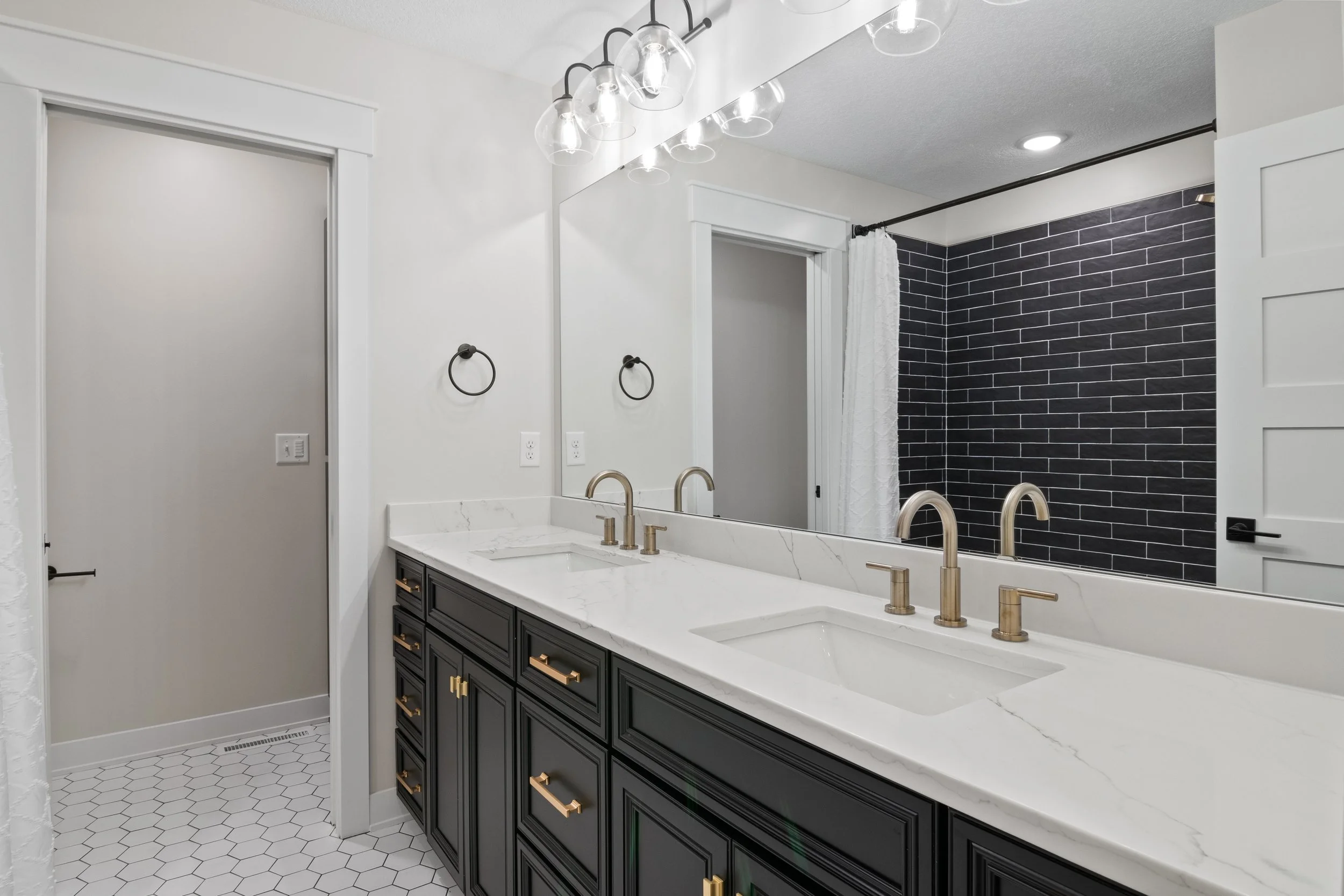 Modern bathroom with white walls, black vanity with gold handles, marble countertop, two gold faucets, large mirror, black towel rings, hexagonal tile floor, and a shower with black brick tiles and white curtain.