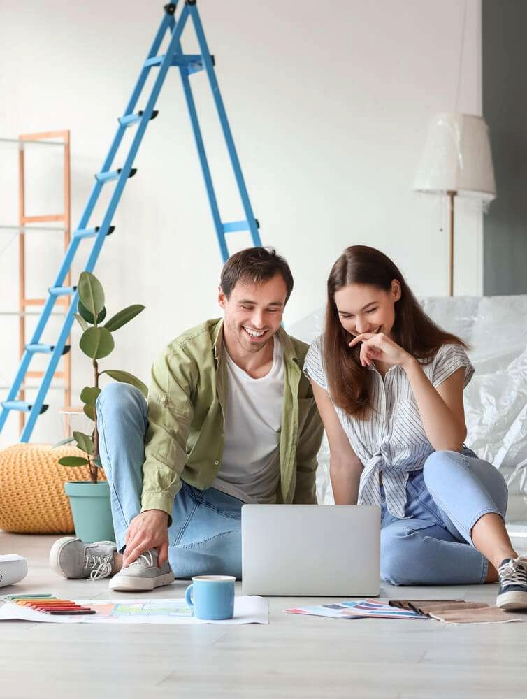 A happy couple sitting on the floor of a room with a laptop, discussing or looking at something on the screen. There are art supplies, a cup, and a plant nearby, with a ladder in the background indicating home improvement or decoration activities.