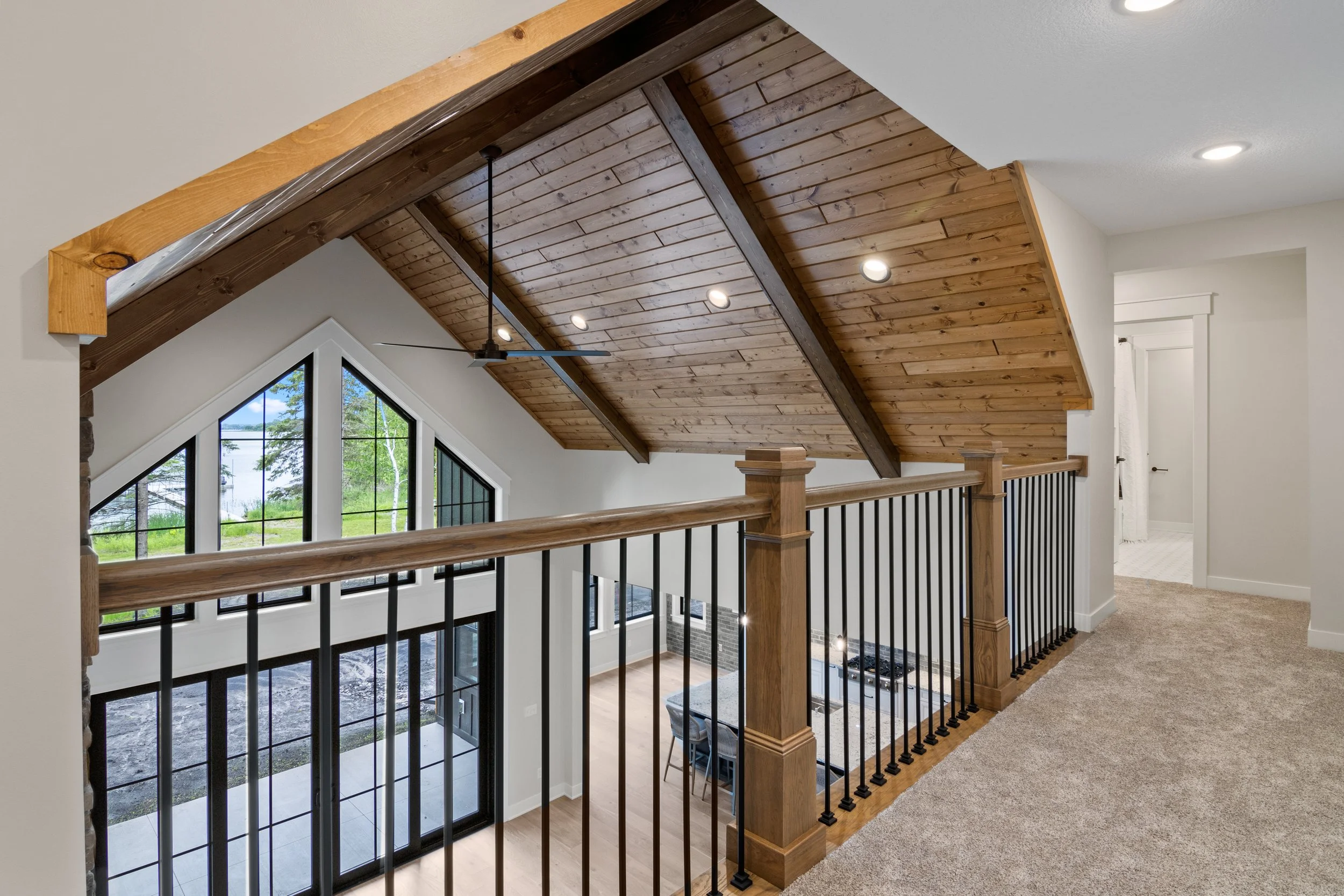 Interior view of a house with a wooden vaulted ceiling, black metal railings, large windows with a view of trees outside, and a partial view of a dining area and kitchen.