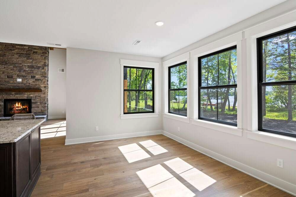 Bright living room with three large windows showing trees outside, hardwood floors, white walls, a fireplace, and a partial view of a kitchen island.