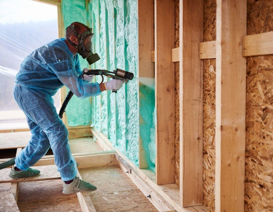 Person wearing protective gear and a helmet using a sprayer to apply spray foam insulation to wall studs in a construction site.