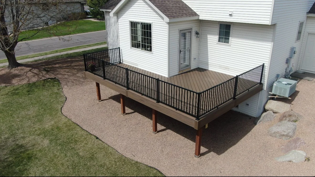 A white house with a newly built wooden deck with black metal railings. The deck has stairs leading to a gravel yard, surrounded by grass and rocks, with a street and trees in the background.