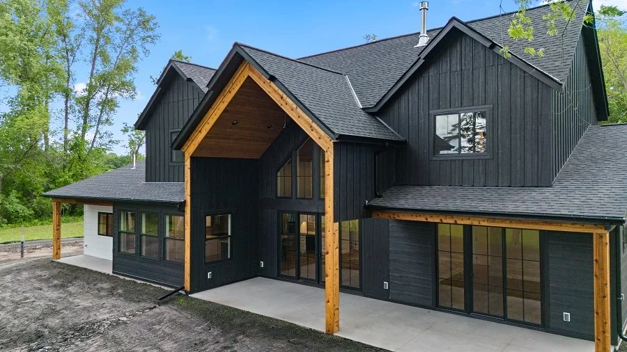 A modern black and wood exterior house with large windows, surrounded by greenery and trees, with a concrete patio area.