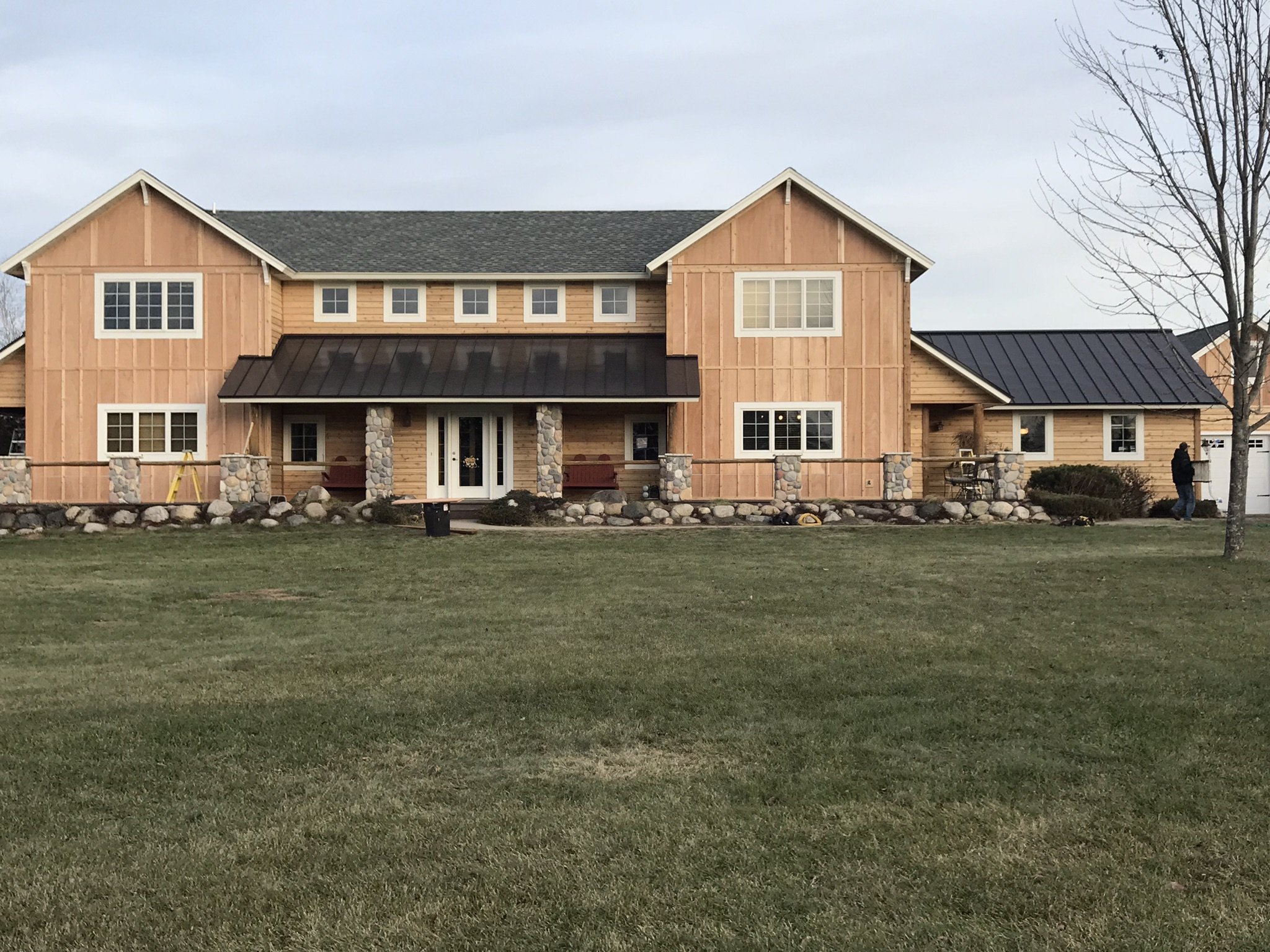 A two-story house under construction with wood siding, stone accents, and a metal roof, surrounded by a grassy lawn.