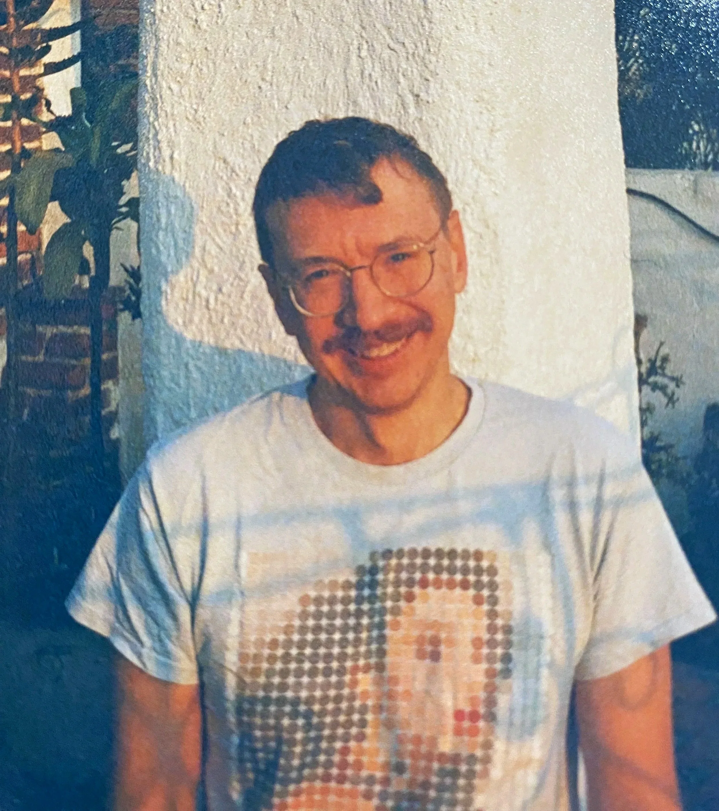 A smiling man with glasses, mustache, and short dark hair, wearing a white t-shirt with a pixelated design of a woman's face.
