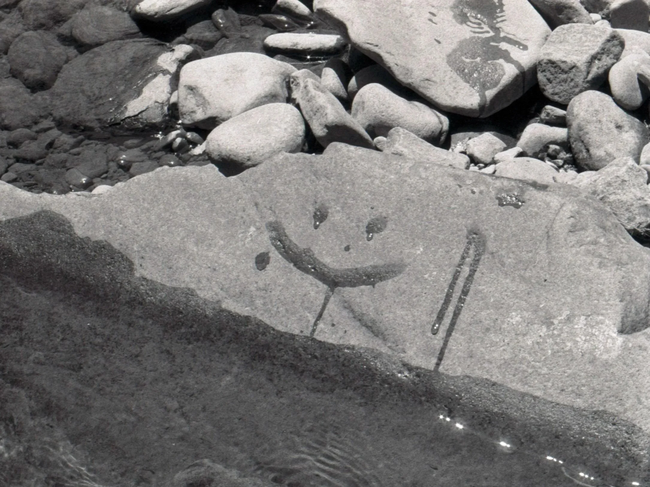 A smiley face is drawn with water on a flat rock among other rocks near a body of water.