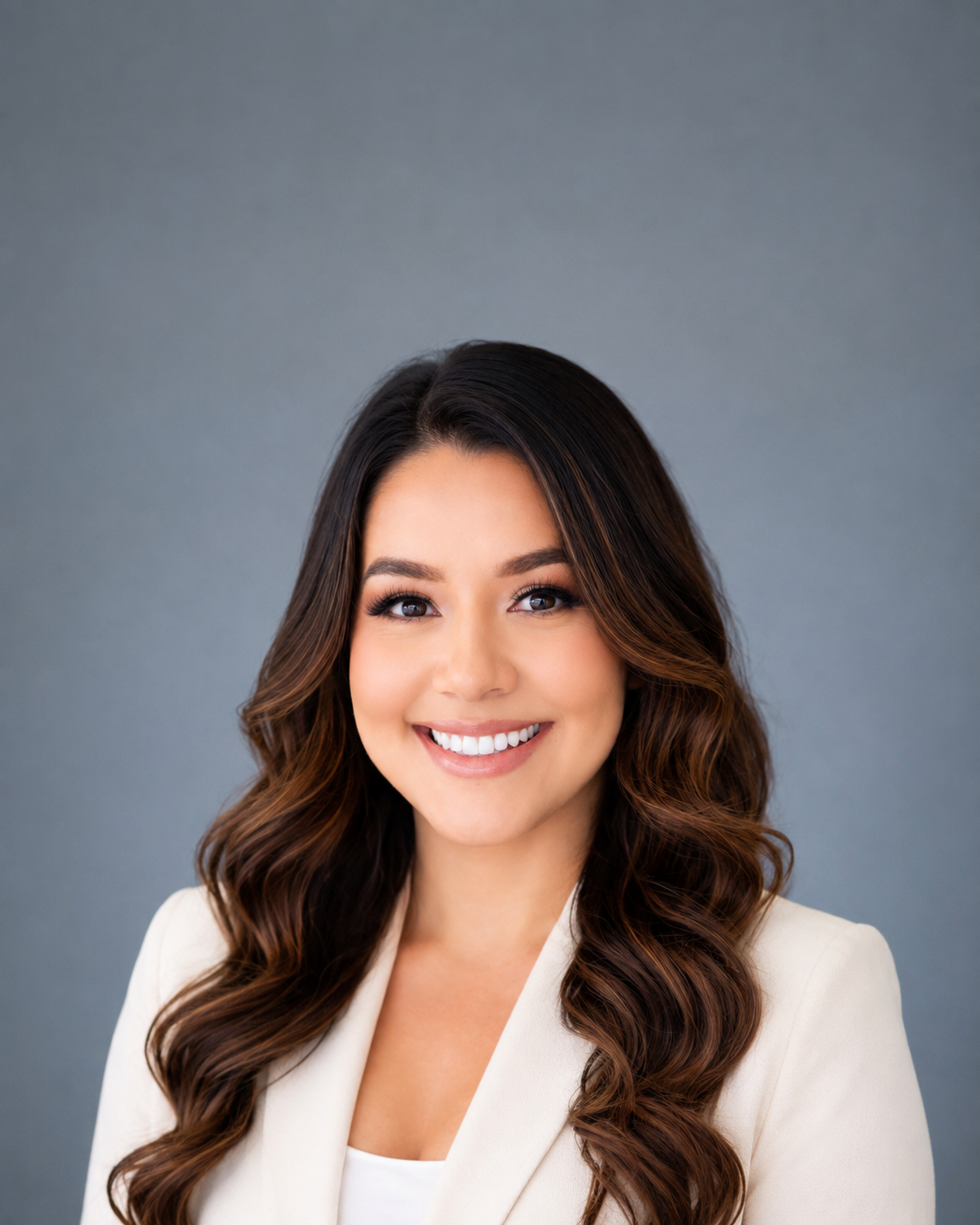 Portrait of a young woman smiling with long wavy brown hair, wearing a white blazer, against a gray background.