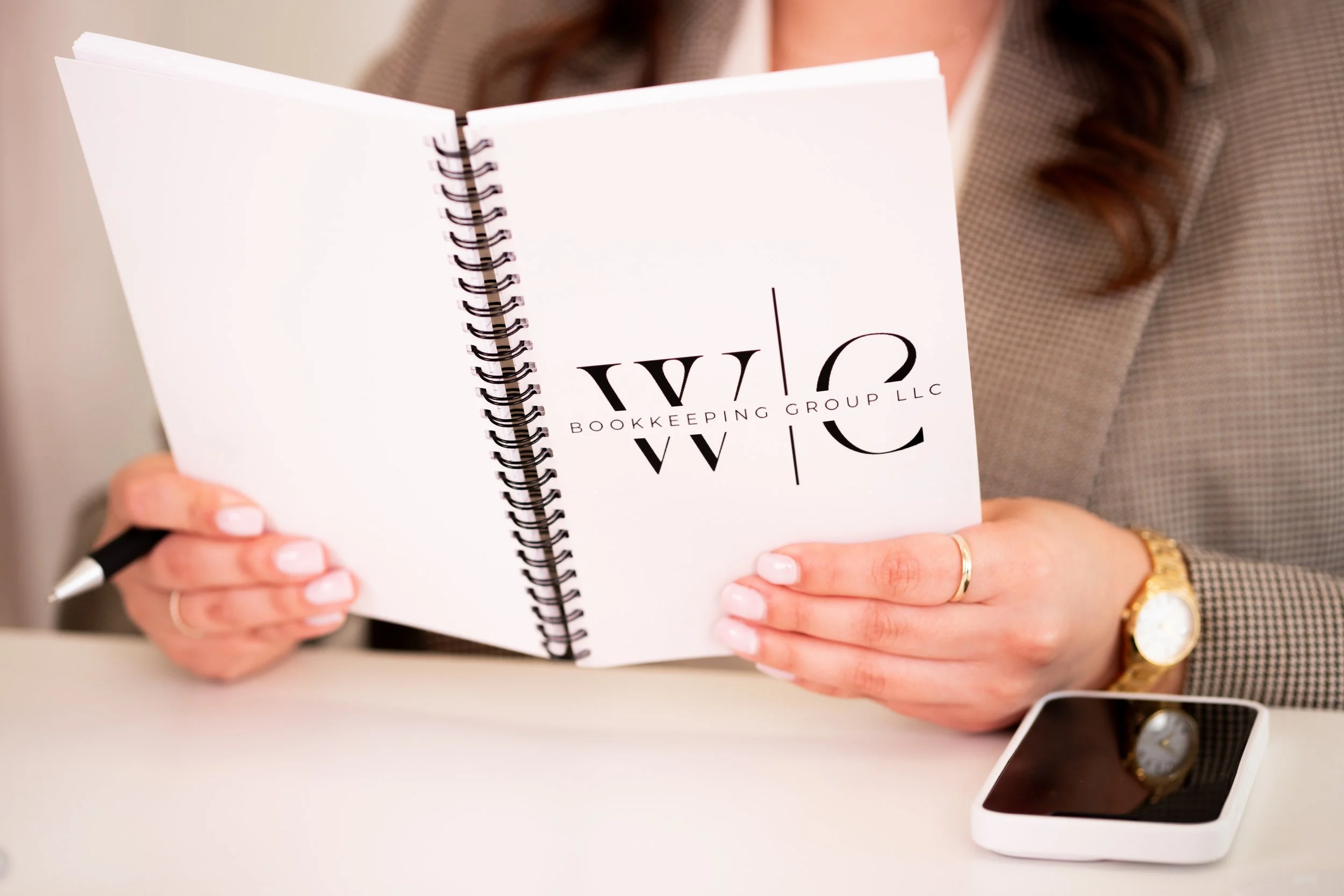 Woman holding a spiral notebook with the words 'Bookkeeping Group LLC' on the cover, sitting at a desk with a pen and a smartphone.