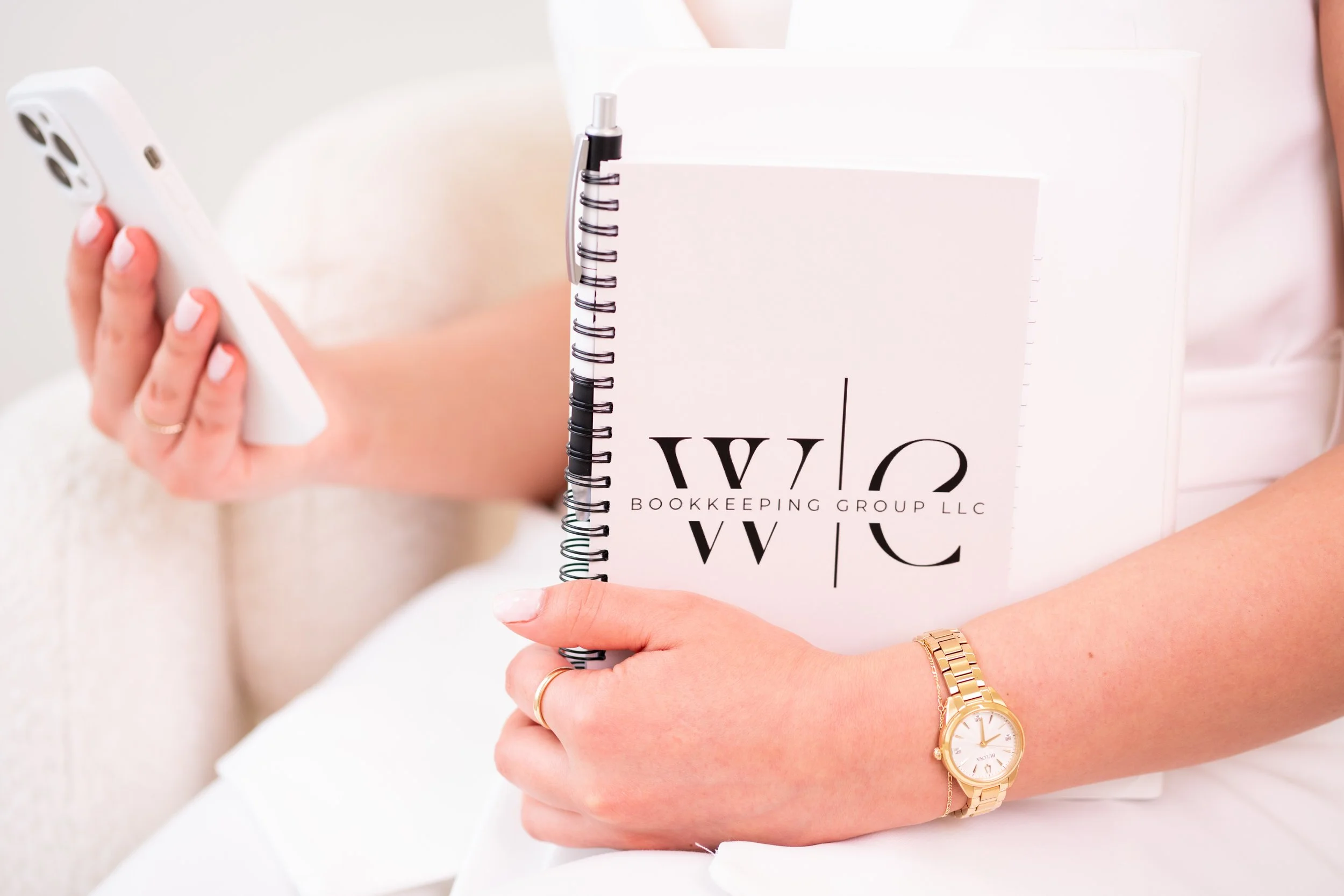 Woman holding a white notebook and a smartphone, wearing a gold wristwatch and a gold bracelet.