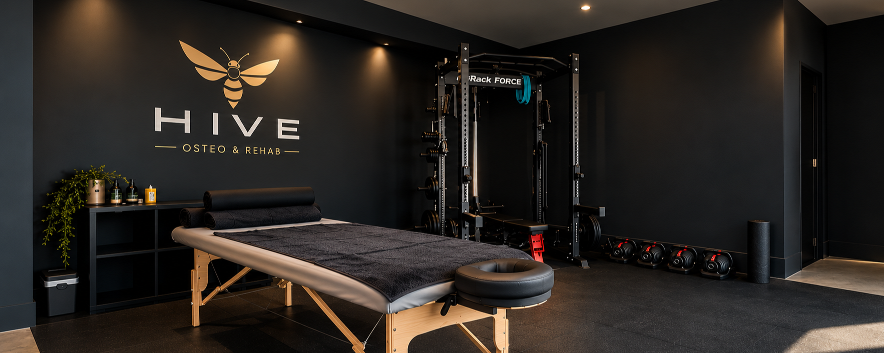 A recovery room in a wellness center with a black wall featuring a bee logo and the words 'HIVE OSTEO & REHAB.' There is a massage or treatment bed with black towels, a black shelf with decorative items and plants, and four kettlebells lined up on the floor. A power rack with weight plates and a bench is also present in the room.