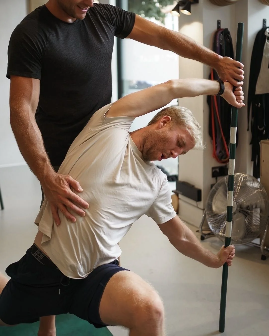A person receiving a physical therapy or chiropractic adjustment from a therapist. The person is kneeling on one knee, holding a pole, and appears to be stretching or exercising, while the therapist supports and guides him.