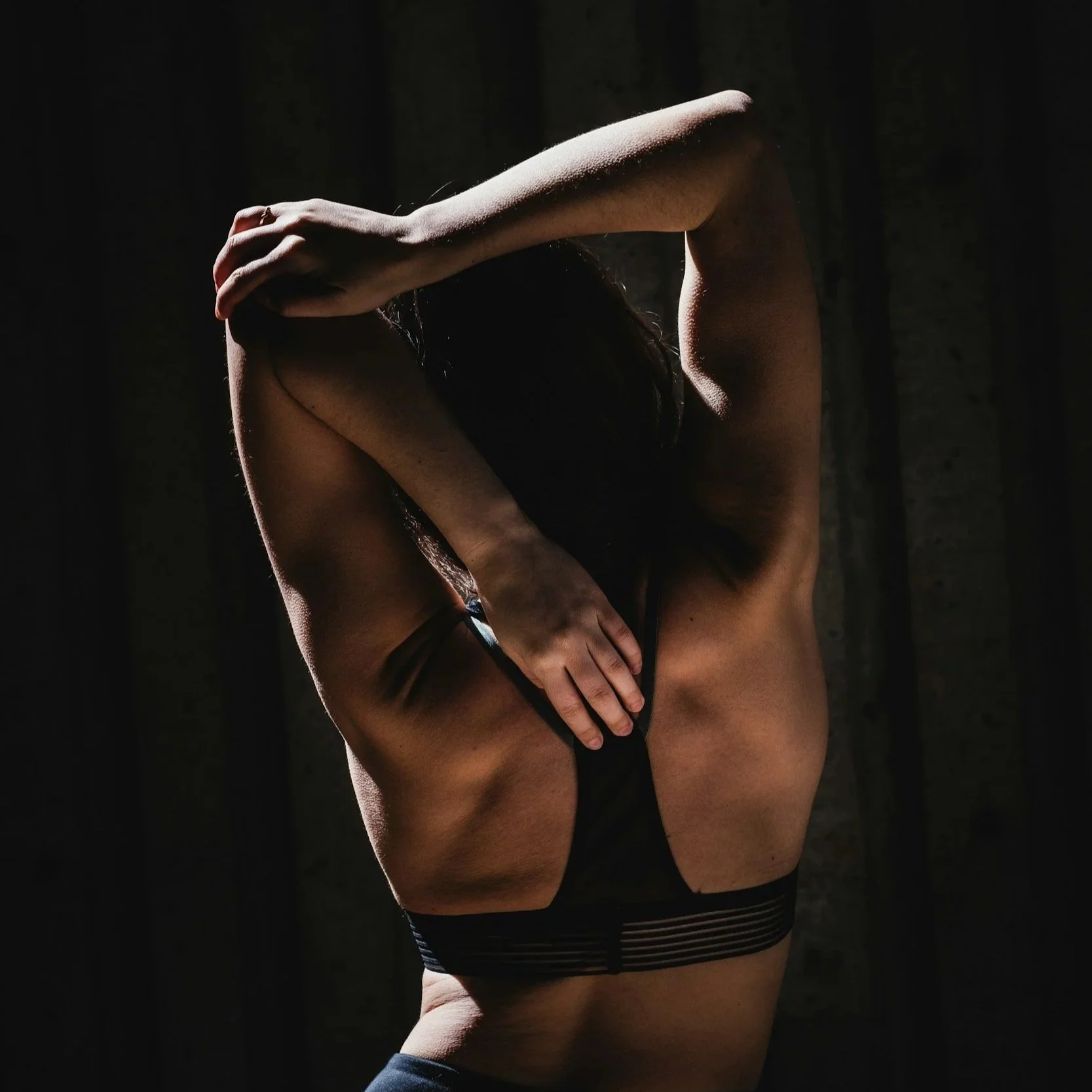 A person with light skin wearing black underwear and a ring on their finger, covering their face with one arm, standing against a dark background with dramatic lighting.