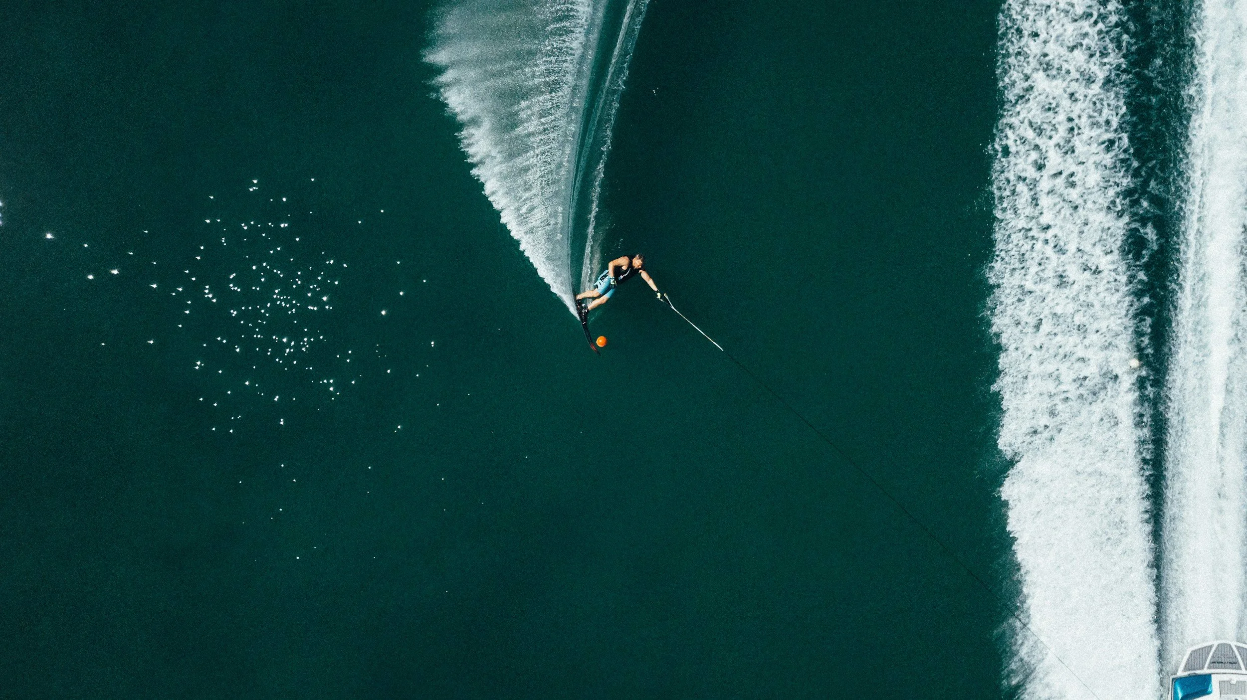 A person waterskiing on a large body of water, holding onto a tow rope with an orange ball at the handle. The water shows ripples and a wake from the boat. The image is taken from above, with a portion of a boat visible at the bottom right corner.