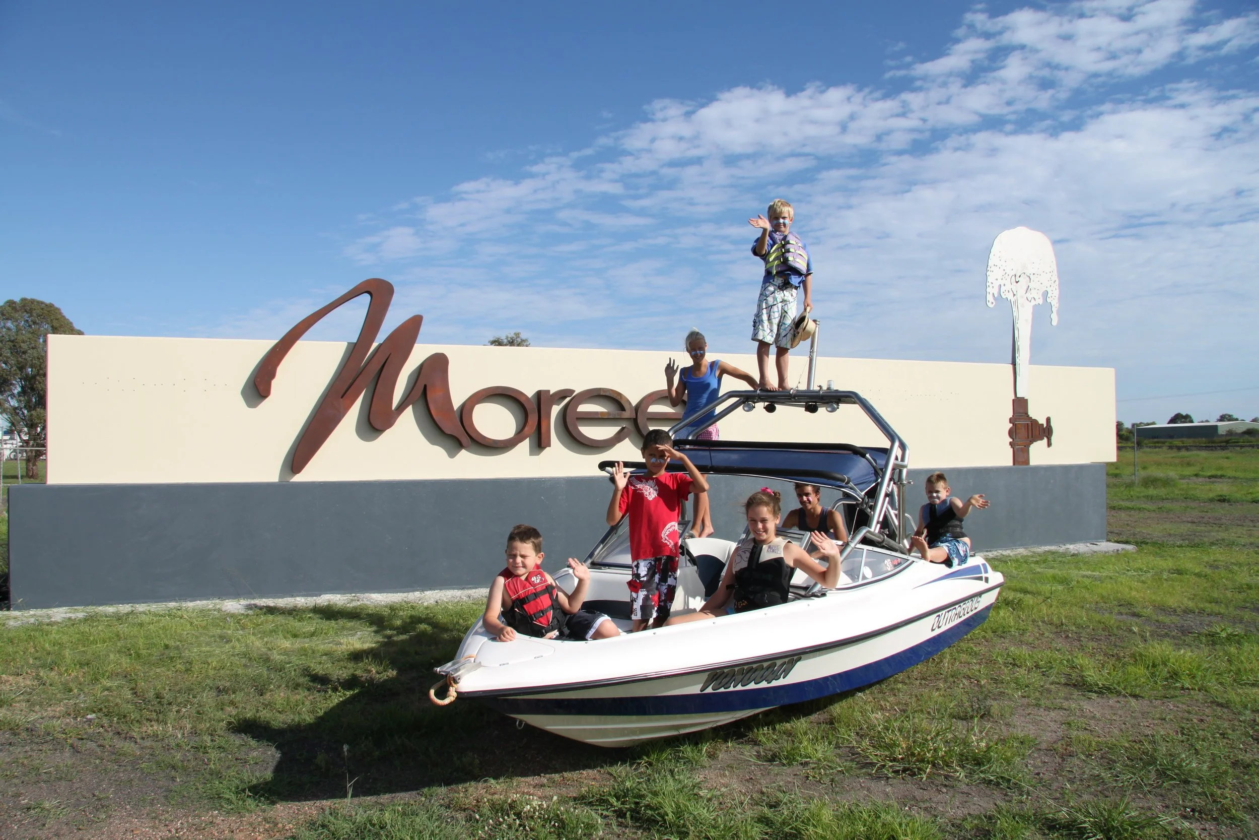 Group of children and a woman riding in a white boat with a black stripe, posing outdoors in front of a sign that reads 'More' with ice cream imagery, on a sunny day with a blue sky and clouds.