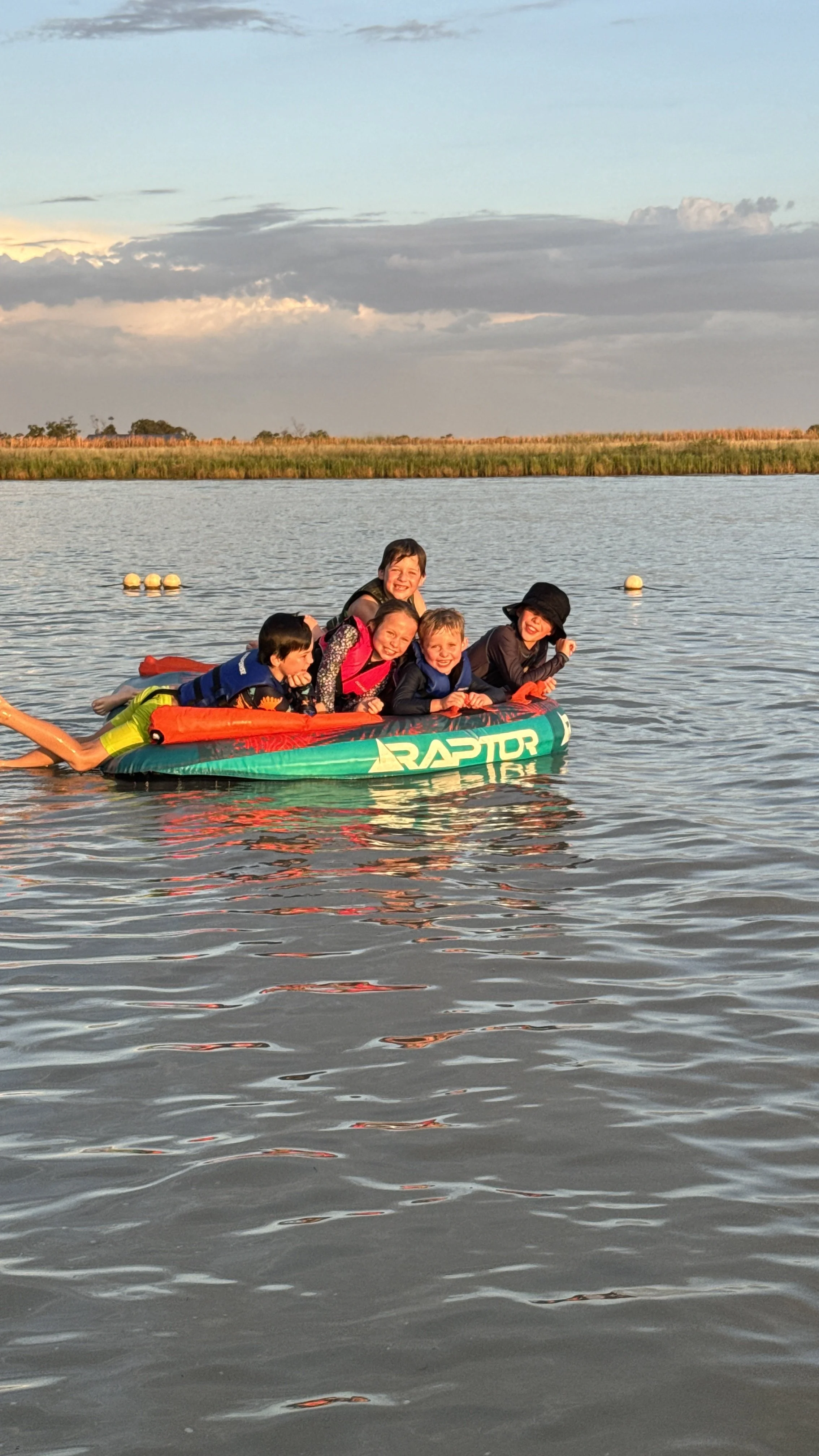 Group of children and a woman smiling on an inflatable boat named 'Raptor' on a body of water, with a grassy shoreline and cloudy sky in the background.