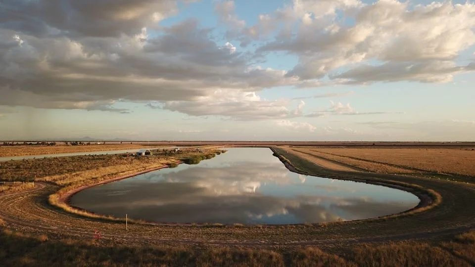 Moree Water Ski Club A large, oval-shaped pond reflecting the cloudy sky surrounded by open farmland under a partly cloudy sky.