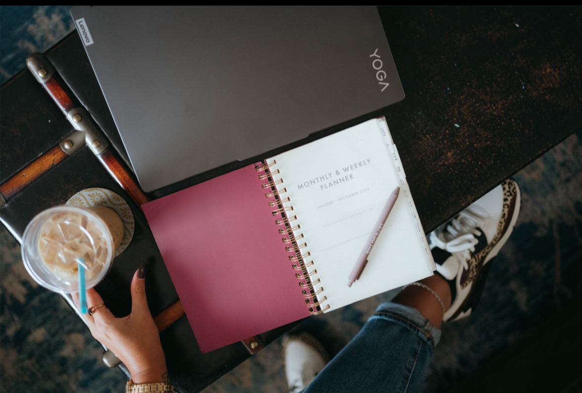 Flat lay of a workspace with a closed gray Lenovo Yoga laptop, a pink and white weekly planner, a silver pen, a hand holding a drink with ice and a blue straw, and part of a person's leg wearing patterned sneakers and a denim pant.