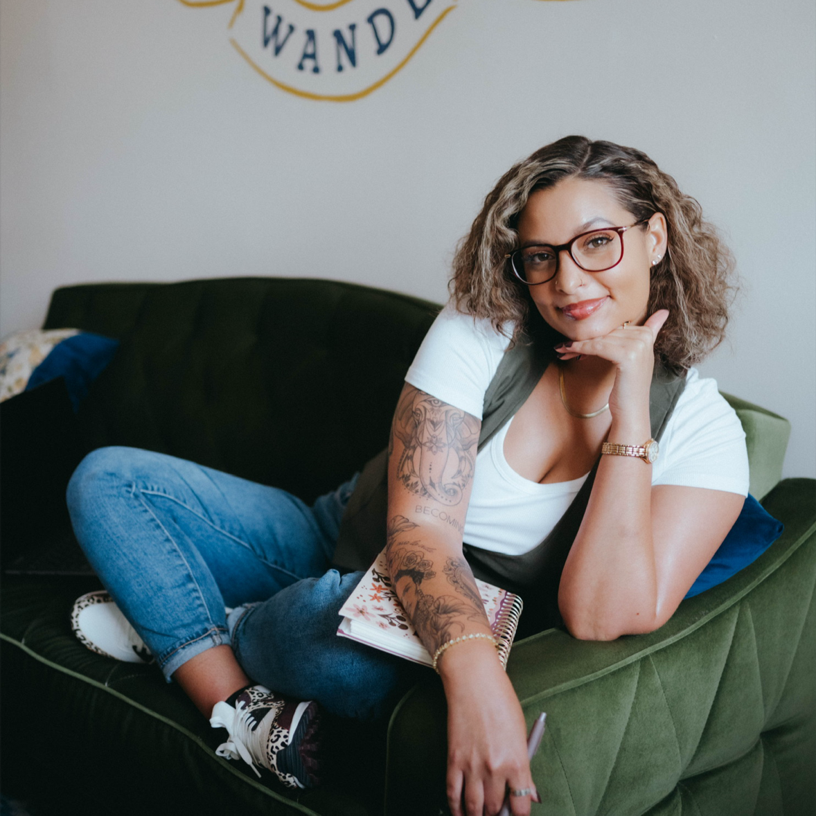 A woman with glasses and tattoos lying on a green velvet couch, holding a notebook and smiling at the camera.