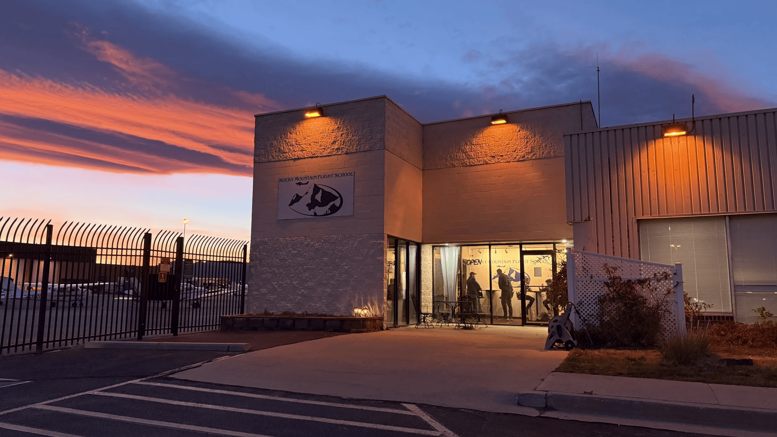 Exterior of Rocky Mountain Flight School building at sunset with a parking lot in front and a fence surrounding the property