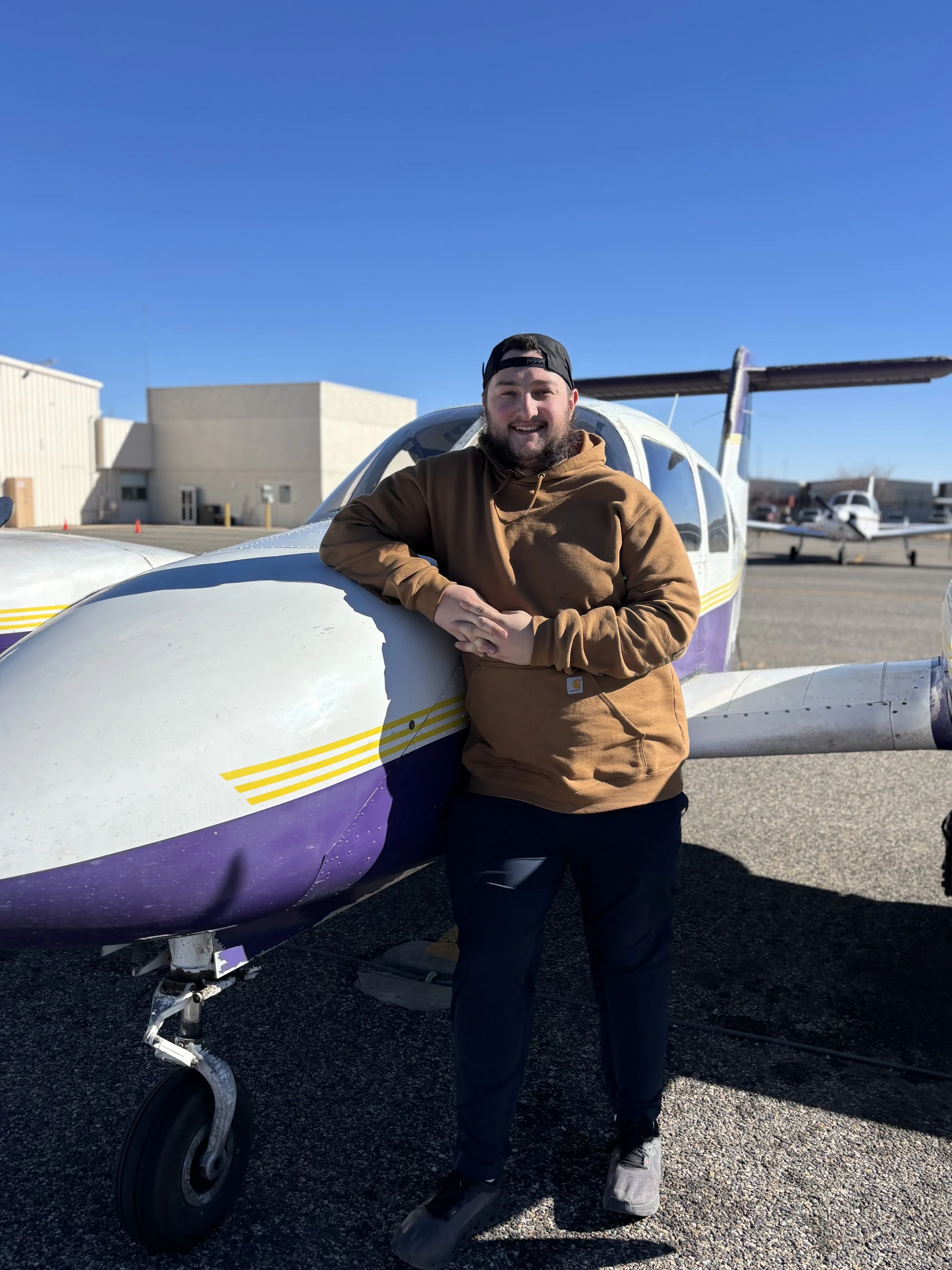 A smiling man in a brown hoodie and black cap standing next to a small purple and white airplane outdoors on a sunny day, with clear blue skies and airport buildings in the background.