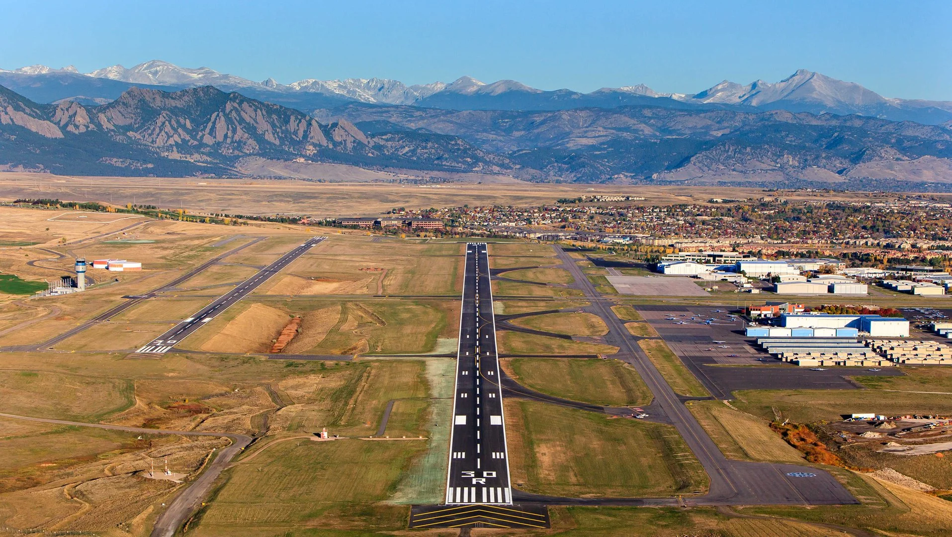 An aerial view of a small airport with a runway marked '50' and 'NORD,' surrounded by fields, industrial buildings, a control tower, and a residential area, with mountains in the background.