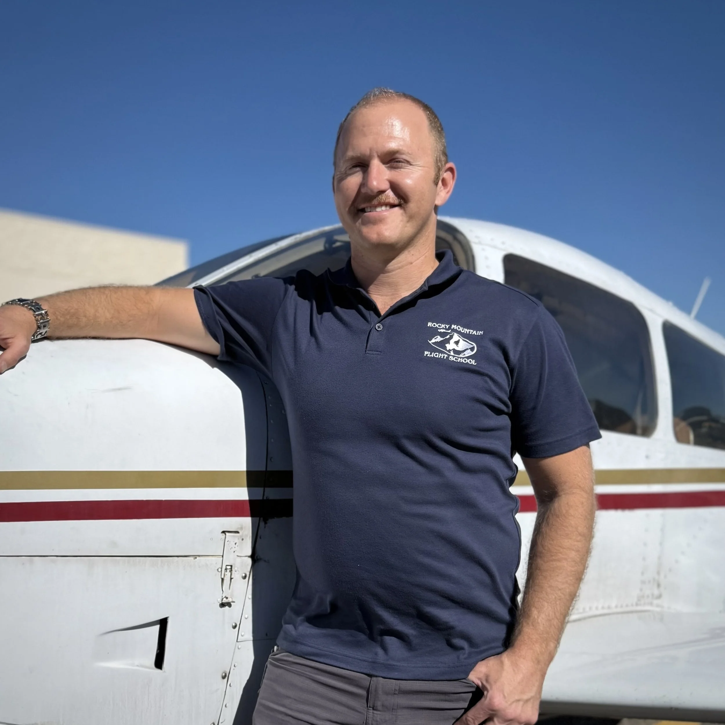 A man with short hair and a goatee, smiling, taking a selfie in front of a small aircraft, wearing a navy blue shirt with 'Rocky Mountain Flight School' logo, with a clear blue sky in the background.