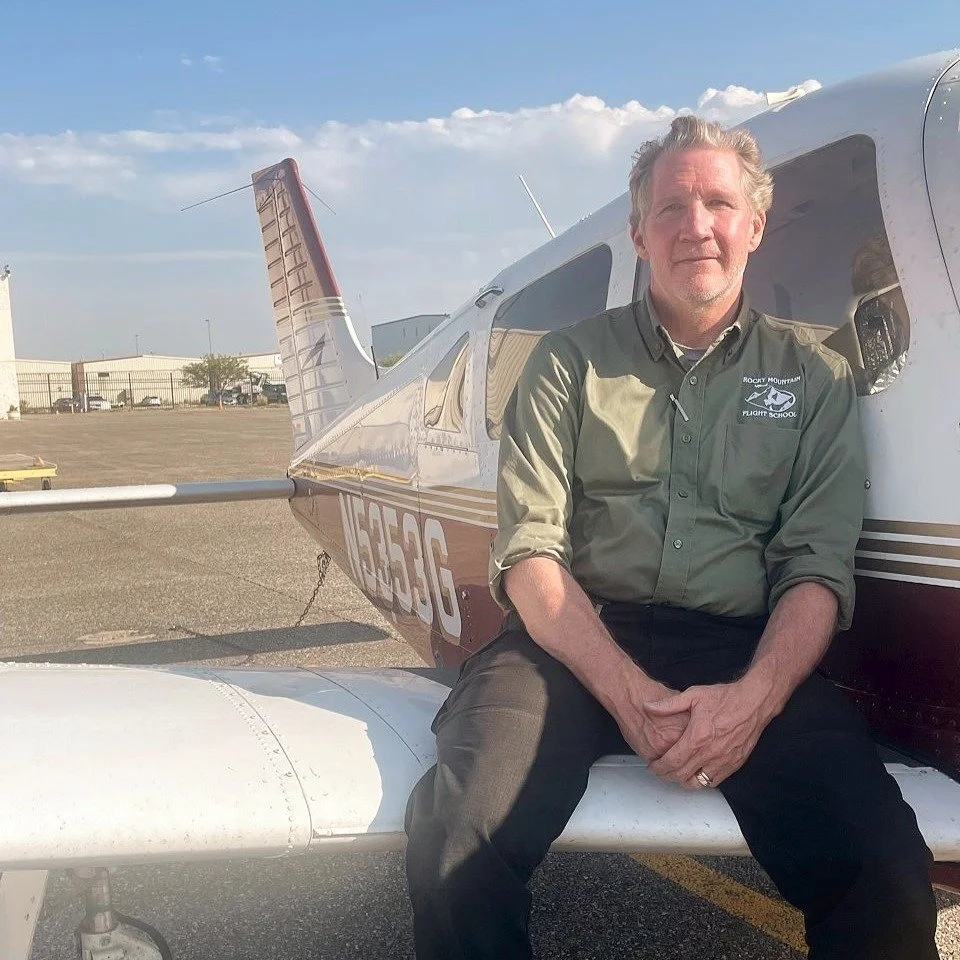 A man sitting on the wing of a small airplane at an airfield, wearing a green flight school shirt, with hangars and a partly cloudy sky in the background.