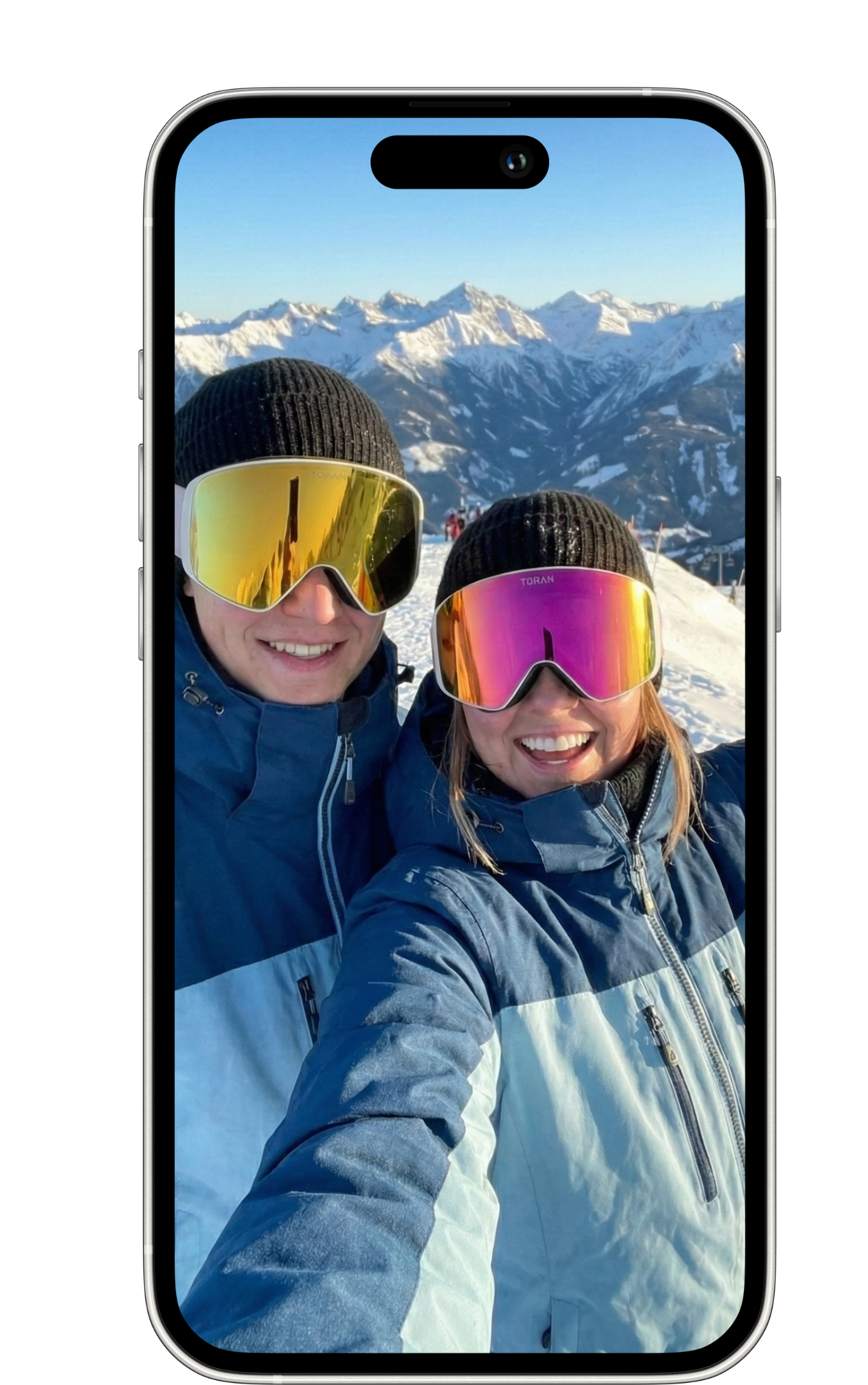 To personer i jakker og skihuer tager selfie på sneklædt bjerglandskab med sneklædte tinder i baggrunden