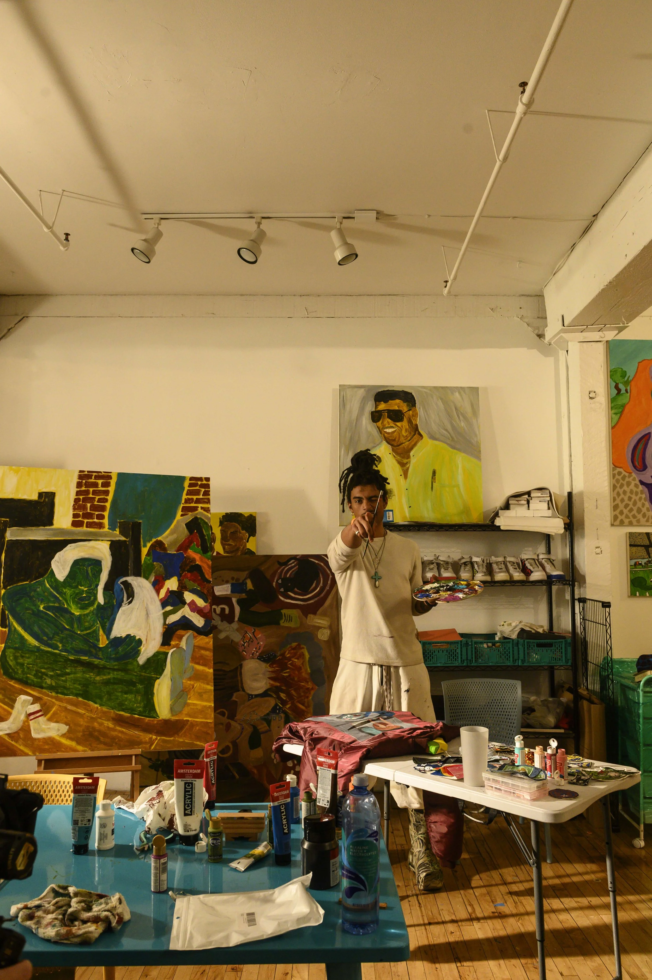 An art studio with a man holding a colorful palette, surrounded by paintings, art supplies on tables, and a wall with various artworks including a portrait and a mural. The room has wooden floors and white walls.