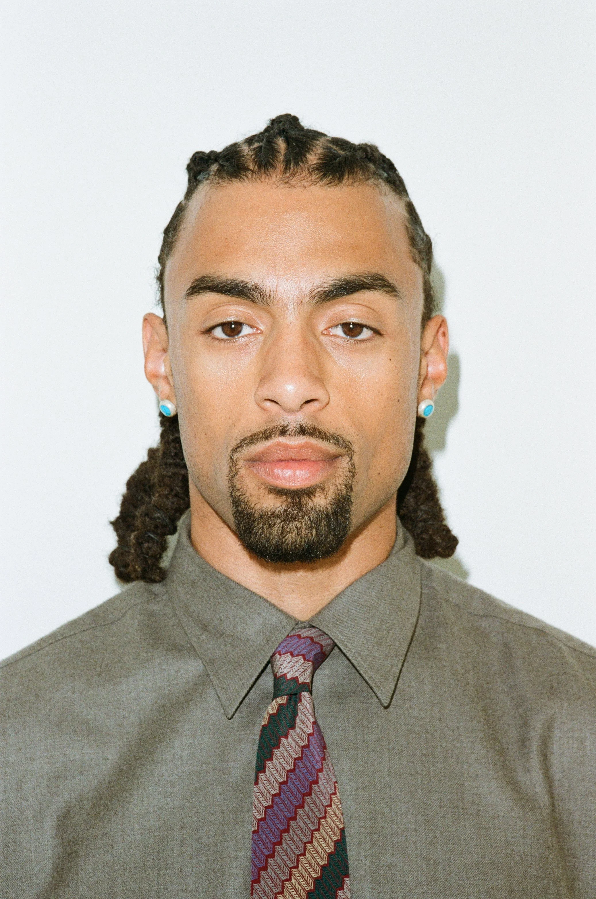 A young man with long braided hair, wearing earrings, a gray shirt, and a patterned tie, looking directly at the camera with a neutral expression.