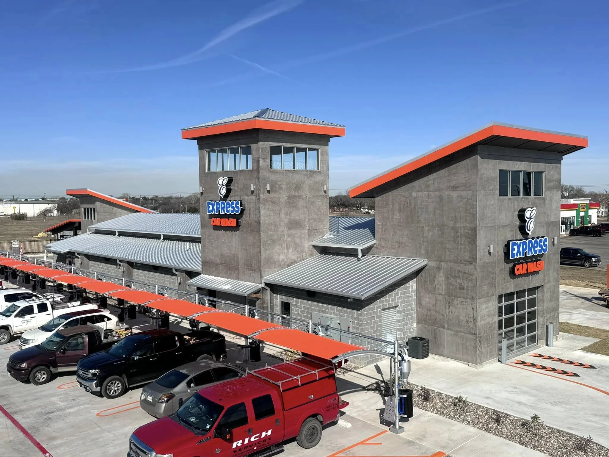 A car wash facility named Express Car Wash with modern concrete and metal roof structures, multiple parked vehicles, and a bright blue sky.