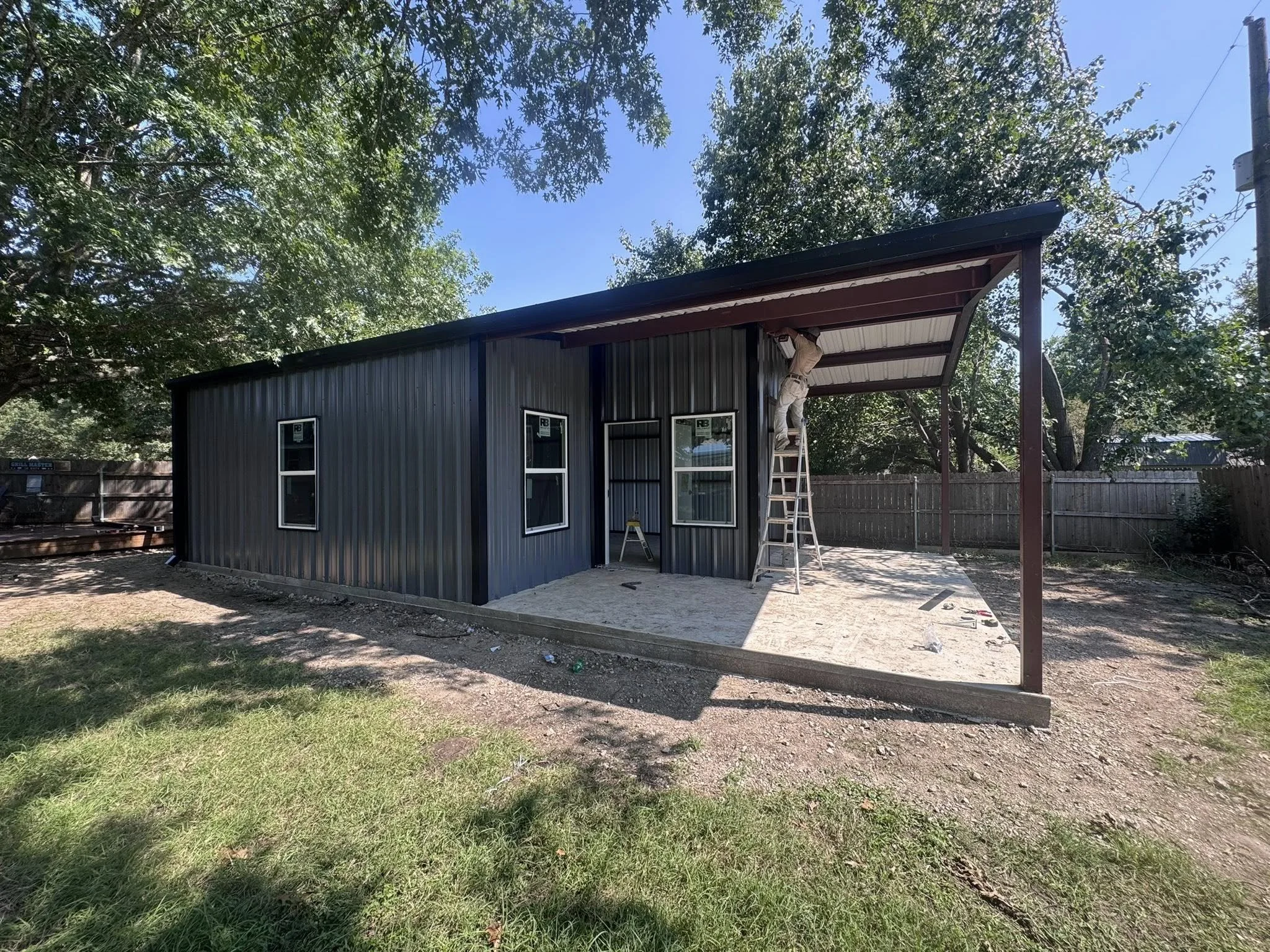 Construction work on a small metal building with a covered porch area, with a worker on a ladder installing or repairing a vent pipe, surrounded by a backyard with a wooden fence and trees.