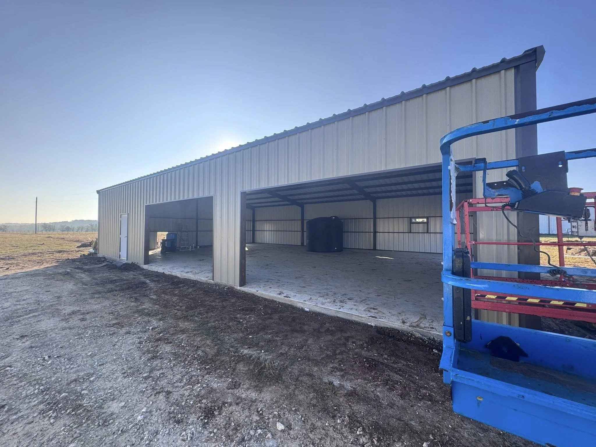 New metal pole barn under construction with a large open front, a smaller closed door, and construction equipment including a blue lift in the foreground, on a gravel and dirt lot with a rural landscape in the background.