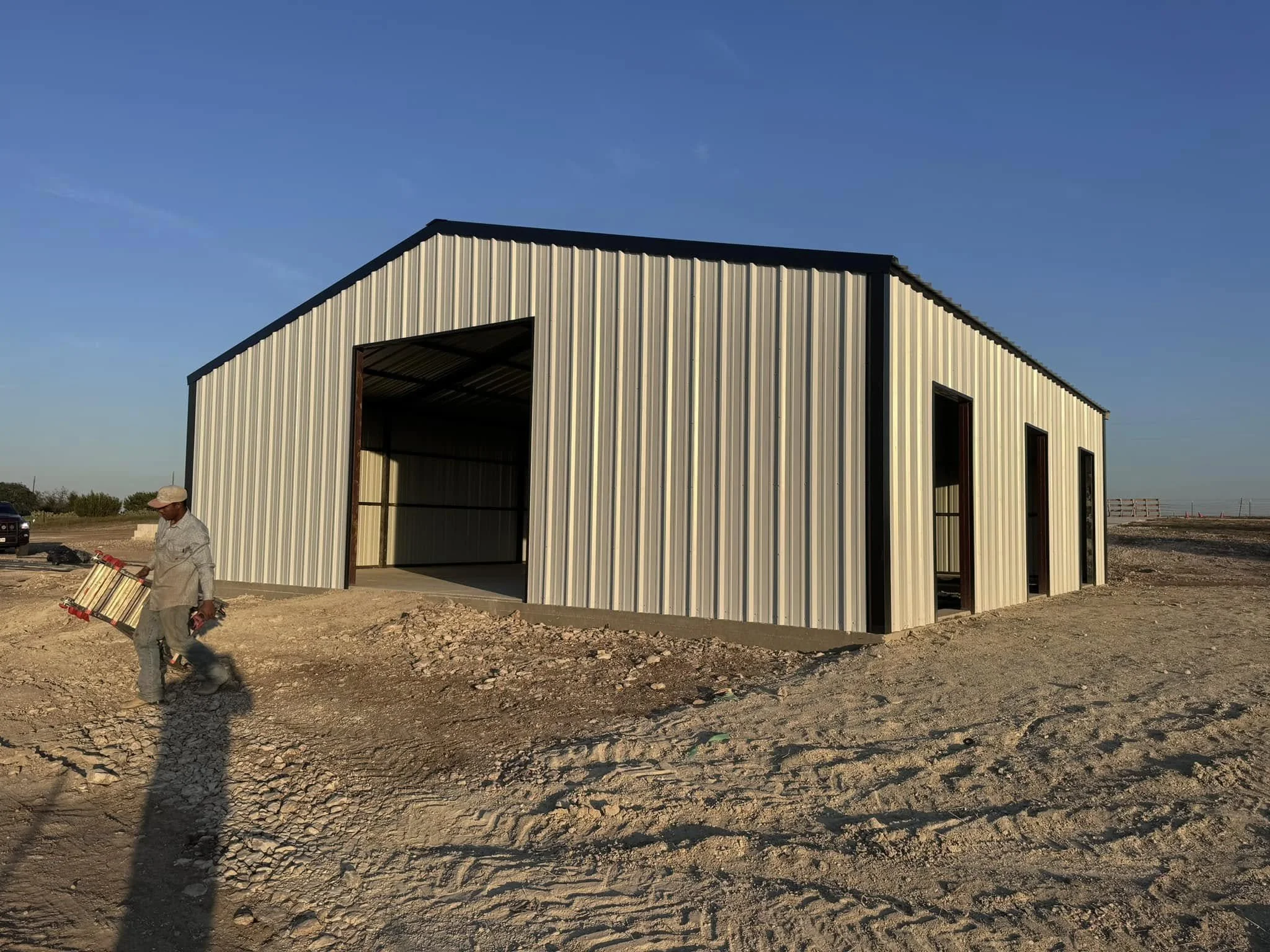 A man walking on dirt ground near a partially constructed metal building with beige siding and black trim, with three door openings, under a clear blue sky.