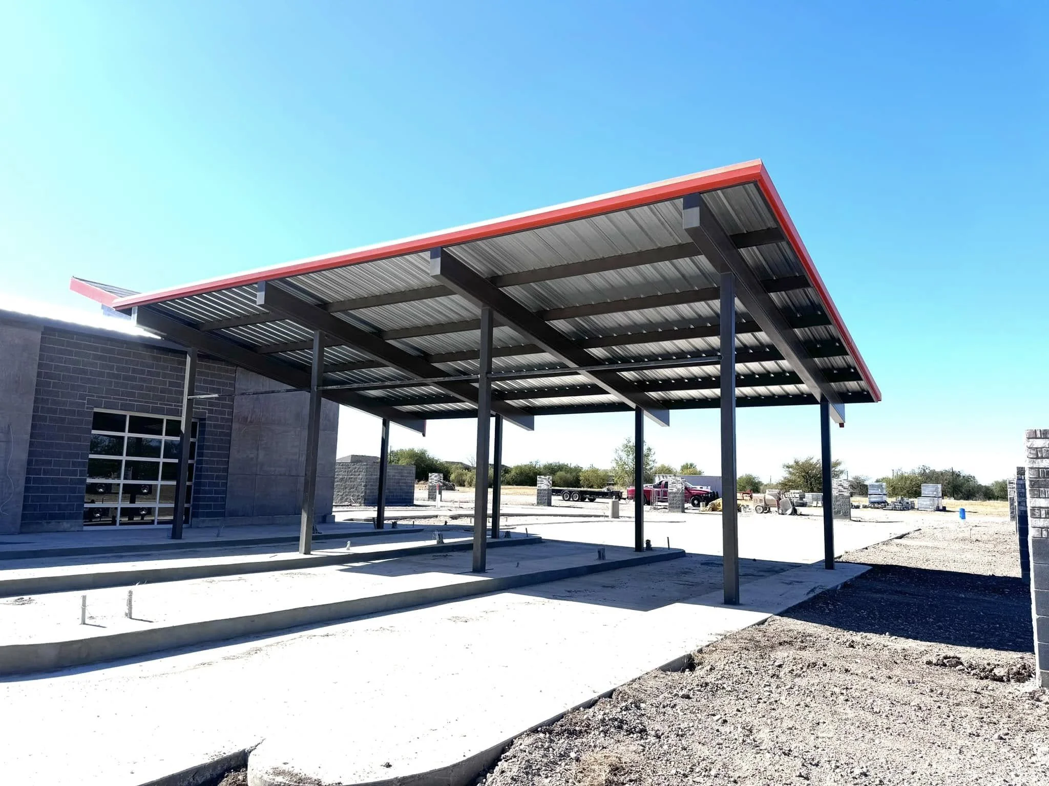 Construction site with a metal carport roof structure in front of a brick building, under a clear blue sky.