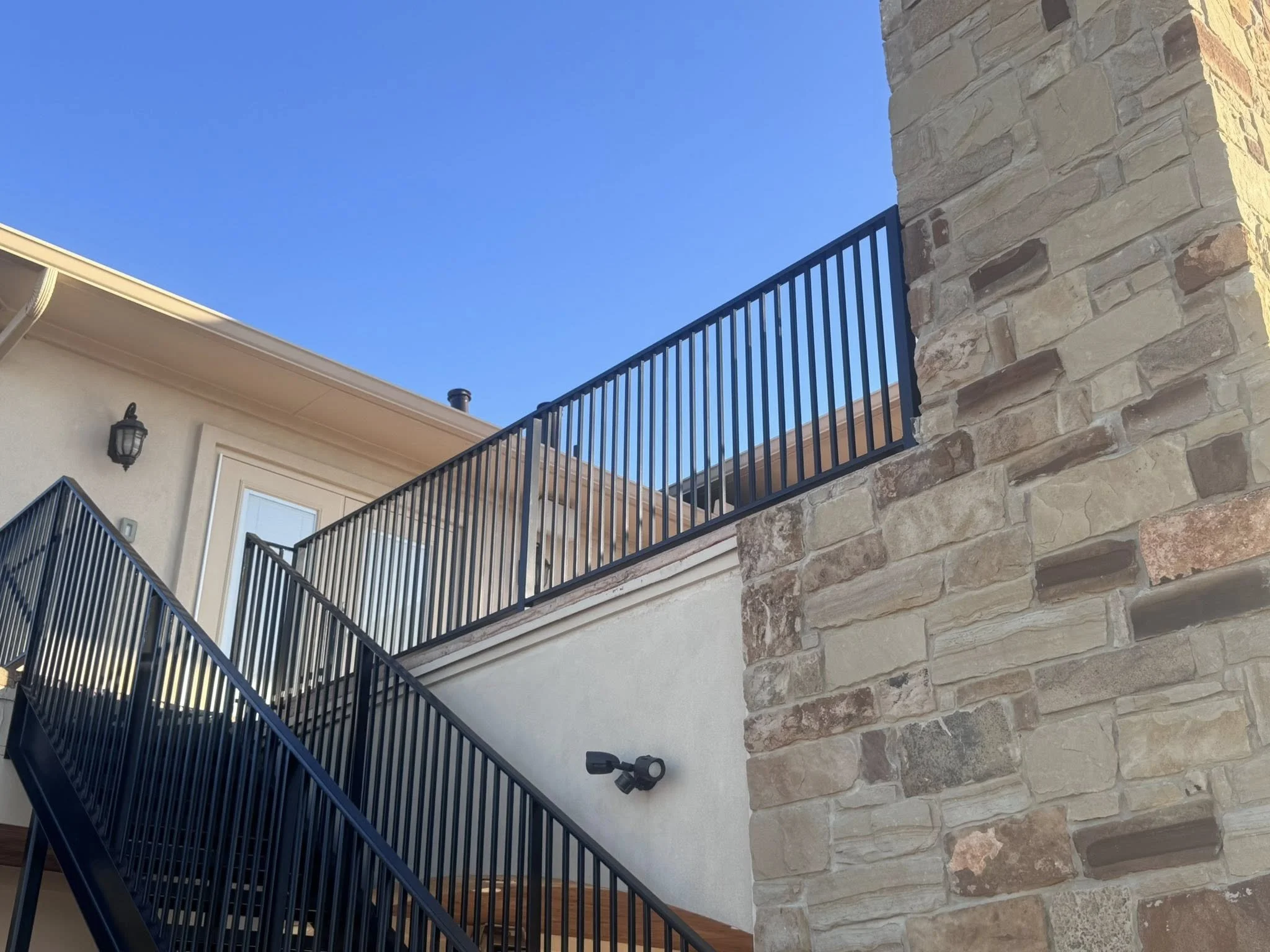 Exterior view of a house showing a staircase with black metal railings, a stone wall, and a door with a light fixture against a clear blue sky.