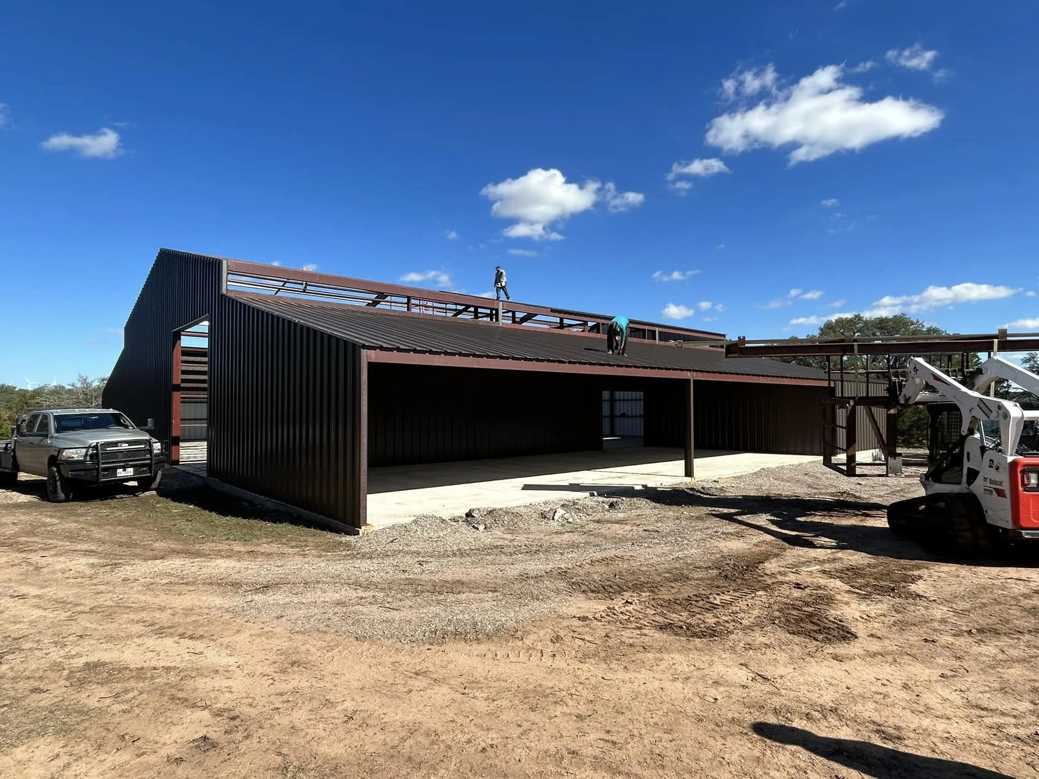 Construction site featuring a modern black building with open garage space, a pickup truck parked on the side, and construction workers on the roof, with a blue sky and scattered clouds overhead.