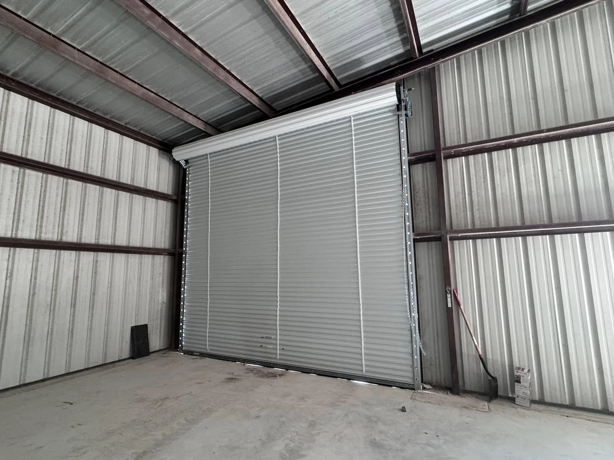 Inside a metal storage unit with a partially open roll-up door, showing construction materials and tools, including a shovel leaning against the wall.