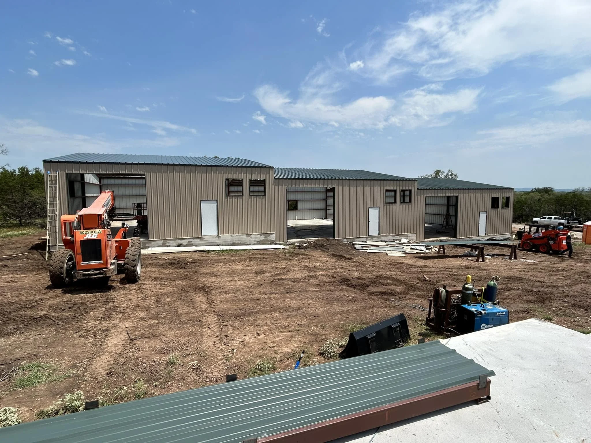 Construction site with metal building in progress, construction equipment, and vehicles on dirt ground under a sunny sky.