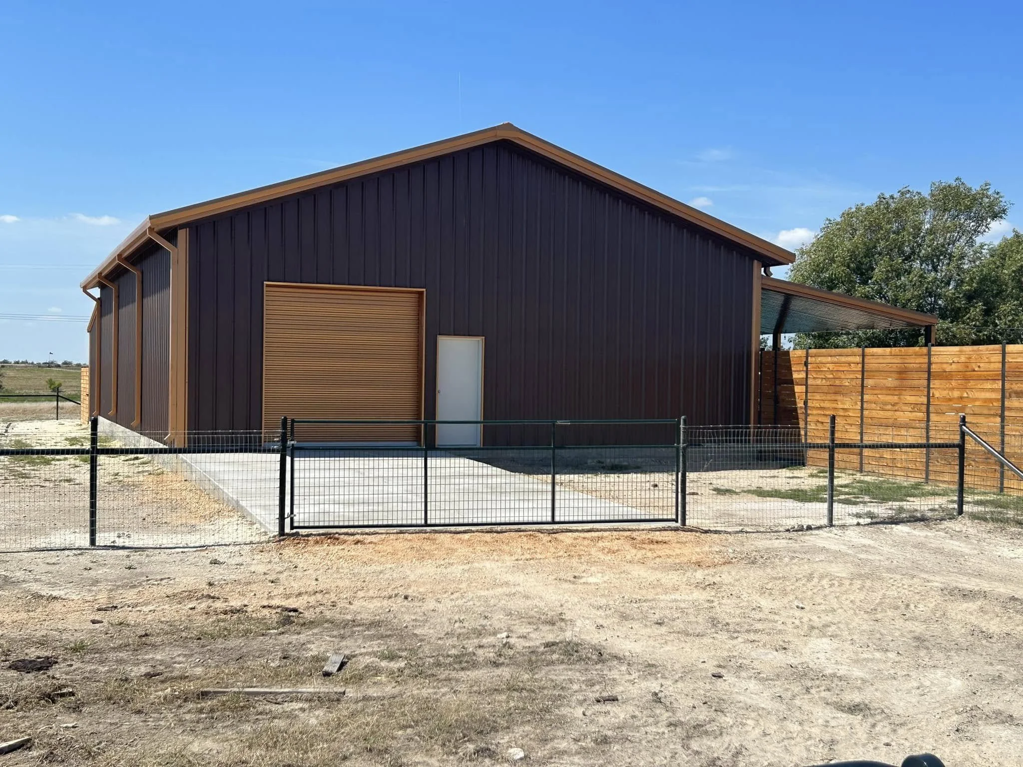 Modern barn with brown siding, a large rolling door, and a small door, surrounded by a black metal fence, on a sunny day with blue sky and trees in the background.