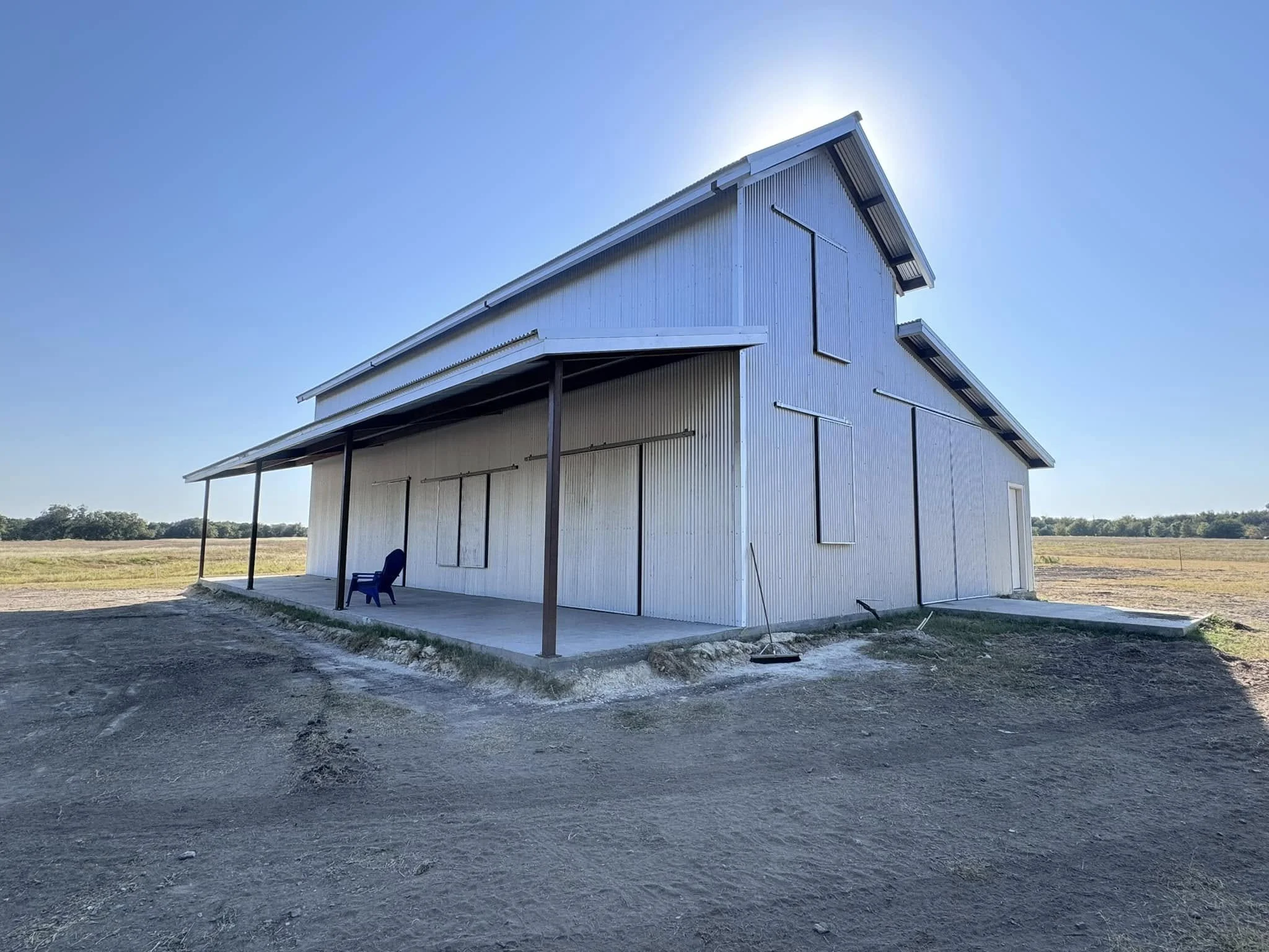 A white metal barn with multiple large boarded-up windows, a small porch with blue chairs, and a sloped roof in a rural area under a clear blue sky.