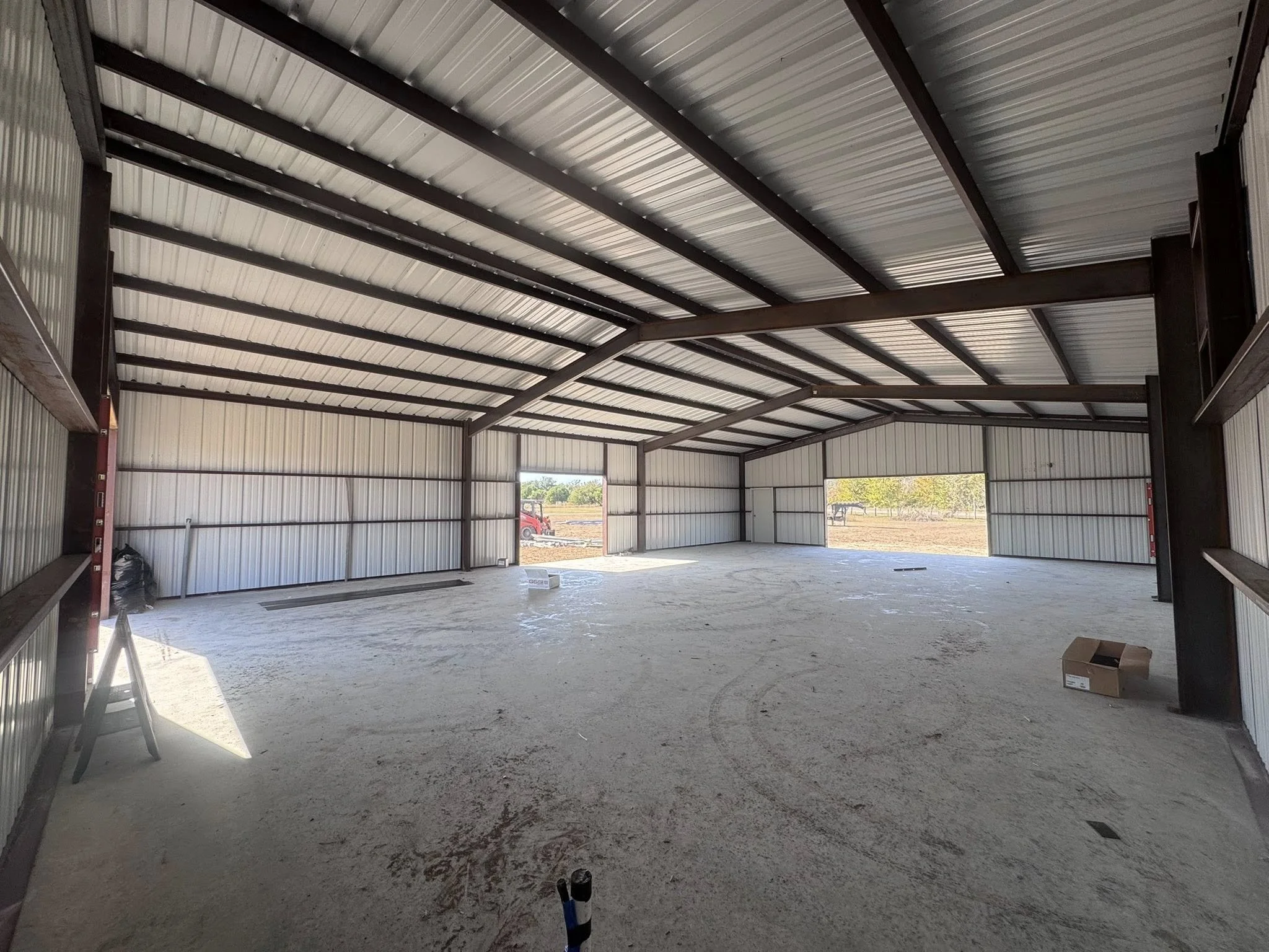 Interior view of an unfinished metal building with a high ceiling, dirt floor, and open garage doors showing outdoor landscape with trees and a vehicle.