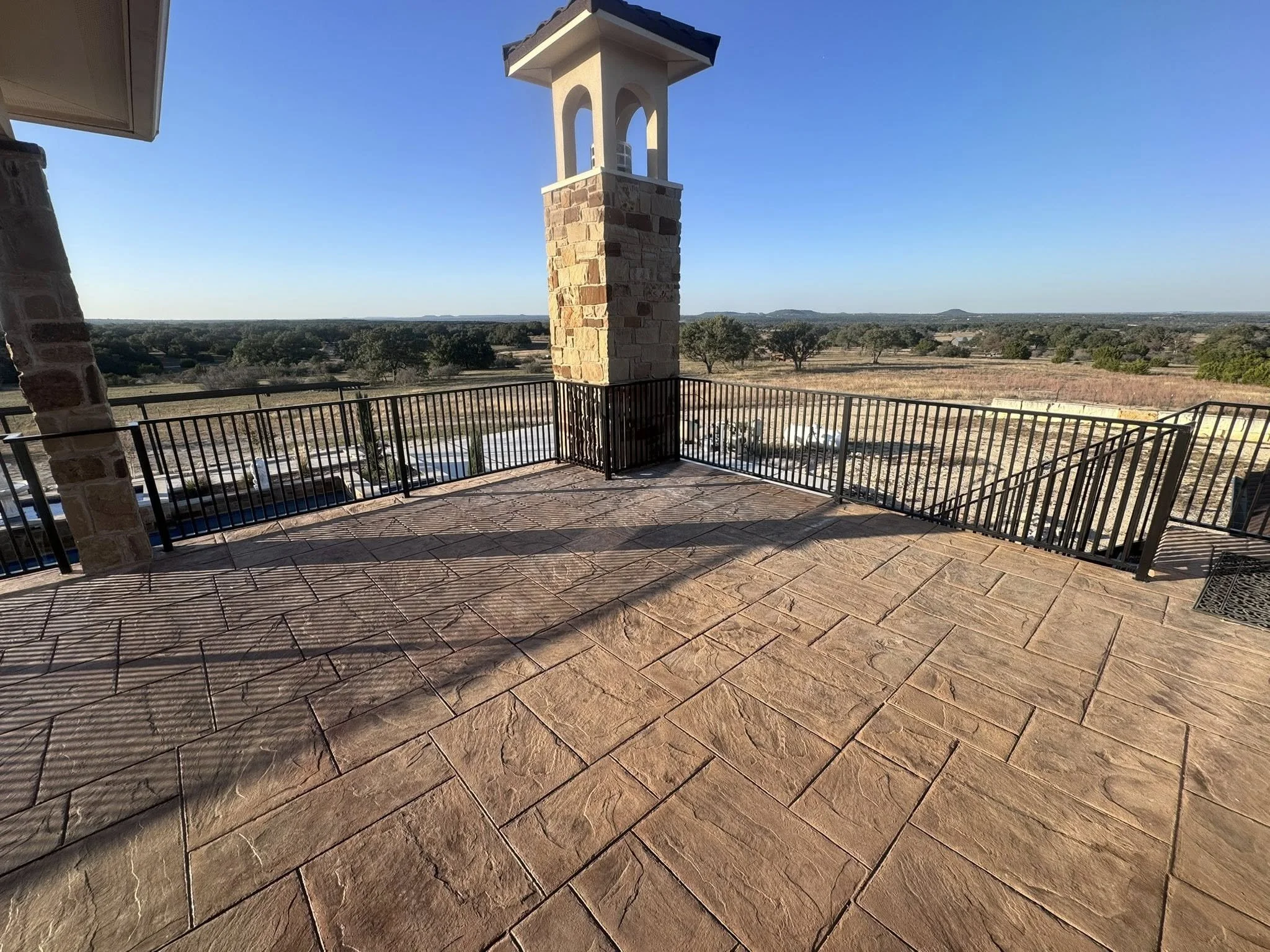 An outdoor balcony with a stone-tiled floor, black metal railing, and a stone and stucco chimney with a bell tower on top, overlooking a rural landscape of open fields and scattered trees under a clear blue sky.