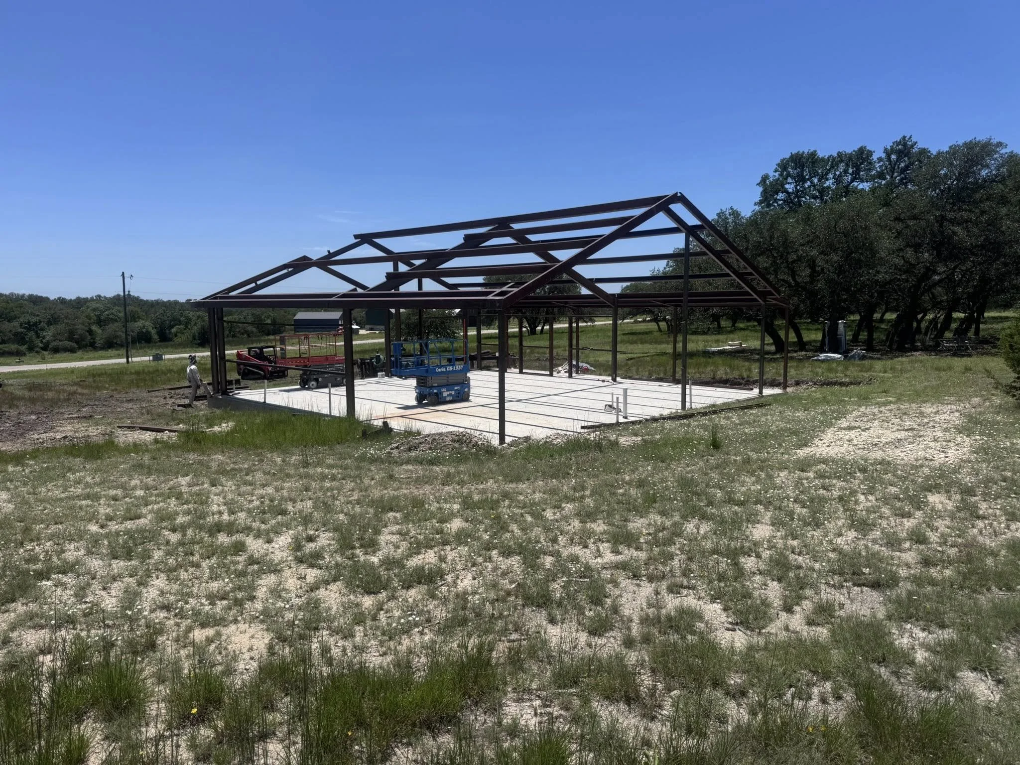 Metal frame structure of a building under construction on a concrete foundation in a rural area with grass and trees.
